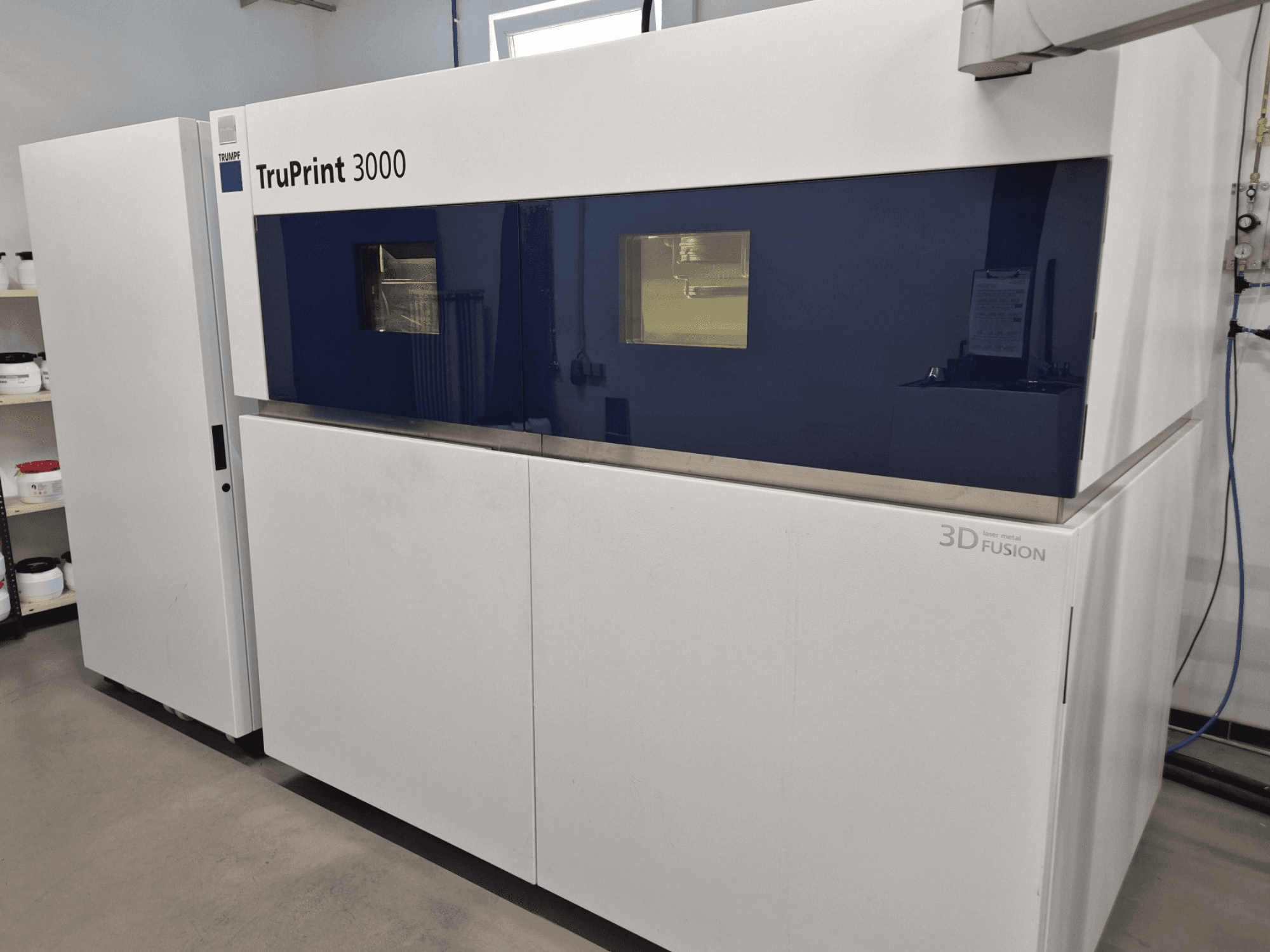 Vista frontal de la impresora 3D TruPrint 3000 para metal, con diseño de paneles azules y tecnología de fusión láser.