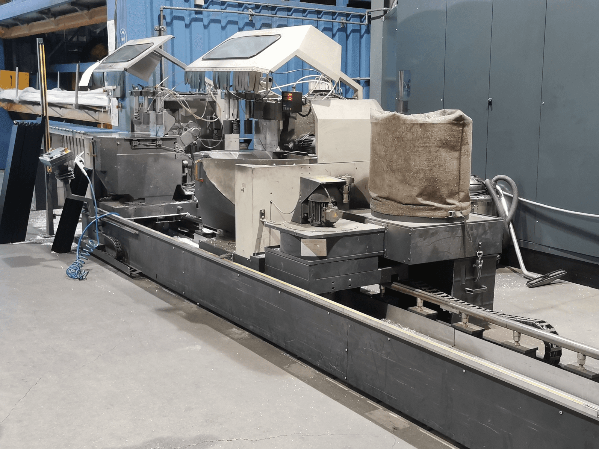 Máquina de procesamiento industrial vista lateralmente, con tolvas, cinta transportadora y panel de control, en un entorno de fábrica.