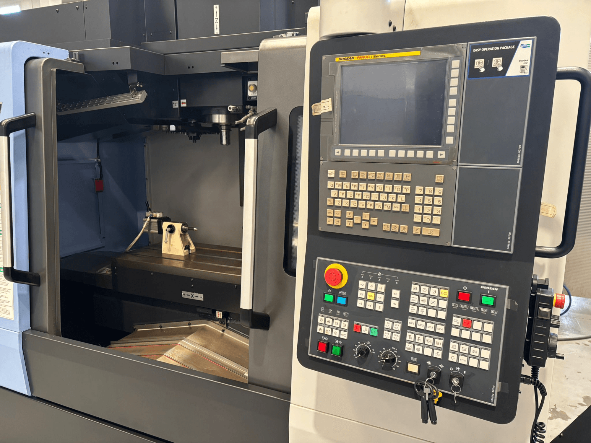 Vista frontal de la máquina CNC Doosan que muestra el panel de control con teclado numérico y botones operativos, con una configuración de pieza de trabajo en el interior.