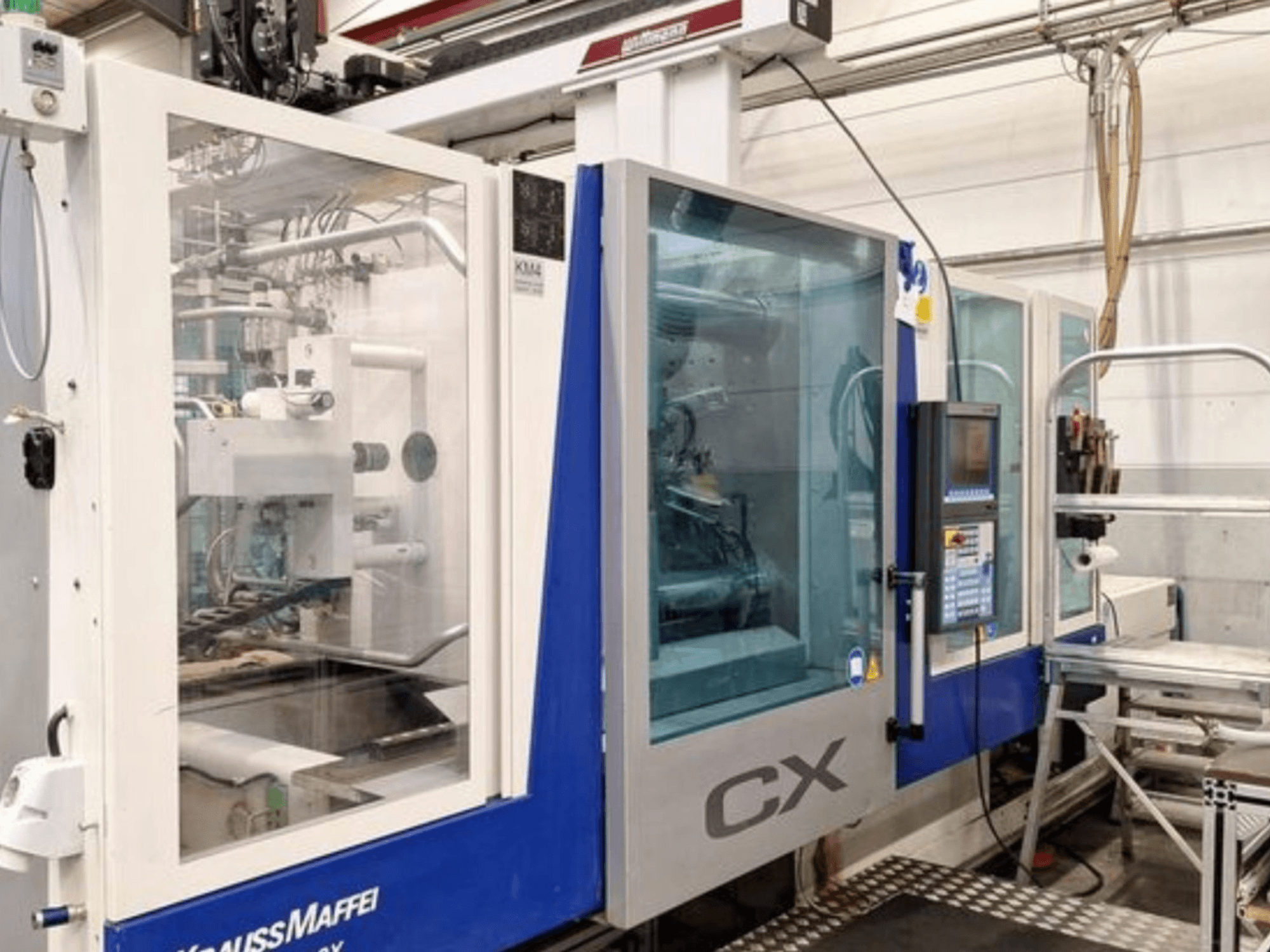 Máquina de moldeo por inyección hidráulica Krauss Maffei 300-1400 CX, vista lateral, en la que se aprecian el panel de control y la carcasa transparente.