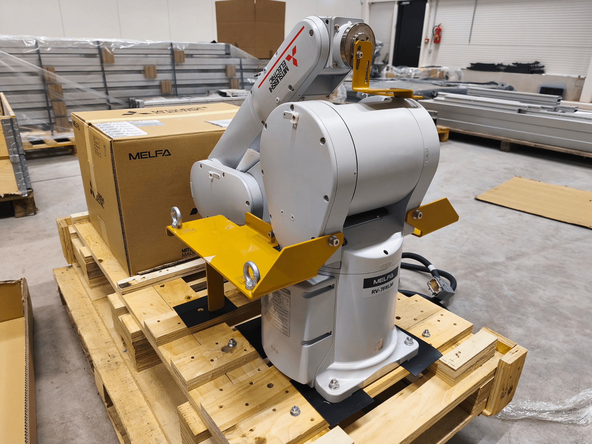 Brazo robótico industrial MELFA RV-7FLM sobre palé de madera, vista frontal, con soportes amarillos y caja de embalaje gris.