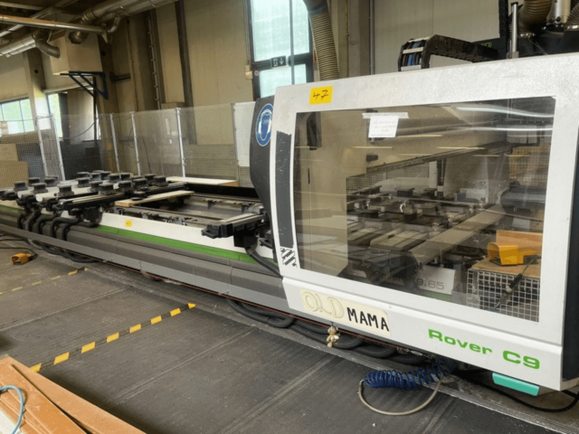 Fresadora CNC BIESSE Rover C 9.65 en un taller, vista lateral mostrando el panel de control y la mesa de trabajo con pinzas.