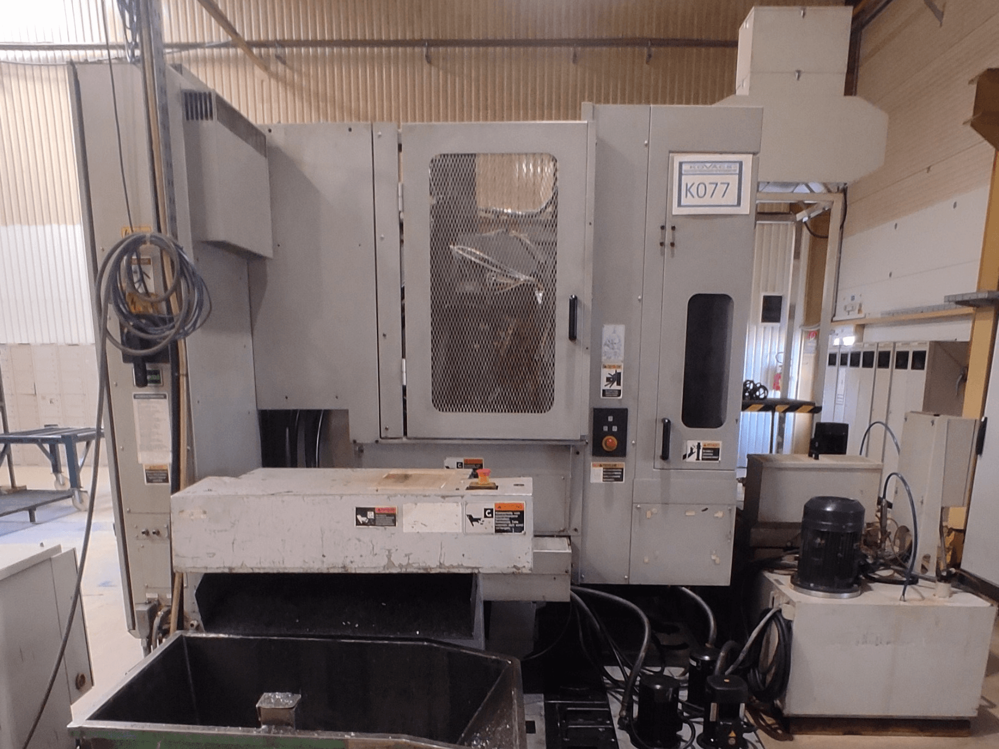 Máquina industrial CNC gris con una gran mesa de trabajo, vista lateral, con panel de control y depósito de refrigerante, modelo K077.