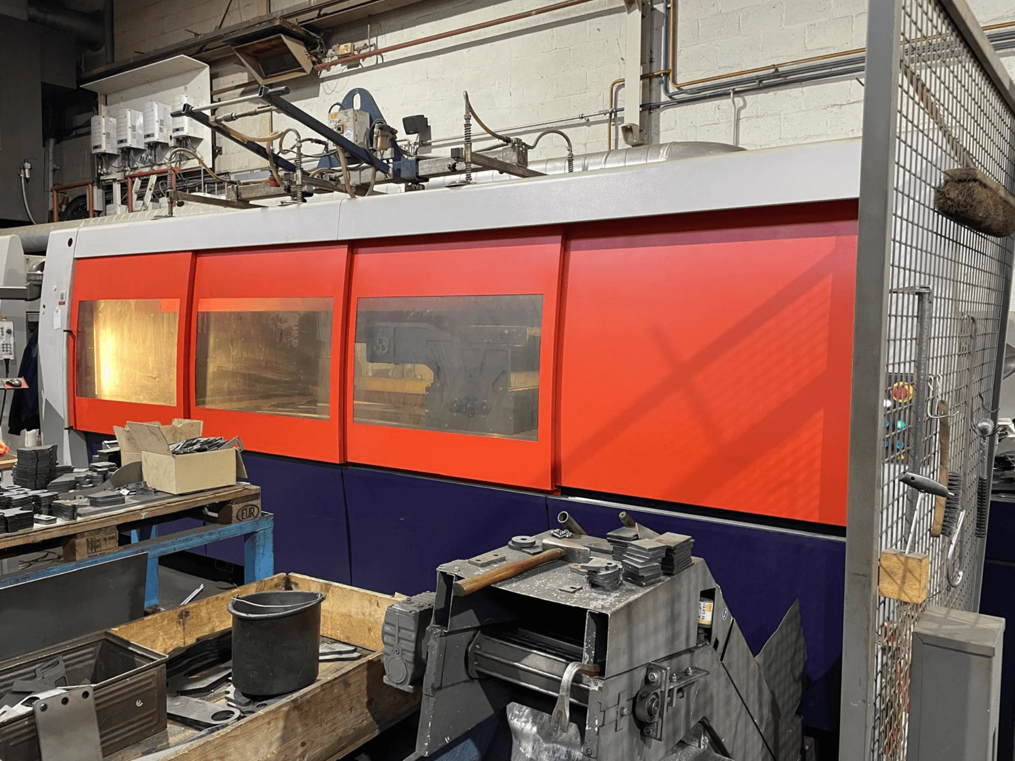 Primer plano de una máquina industrial de corte por láser naranja y blanco, vista lateral, con panel de control y herramientas de taller en primer plano.