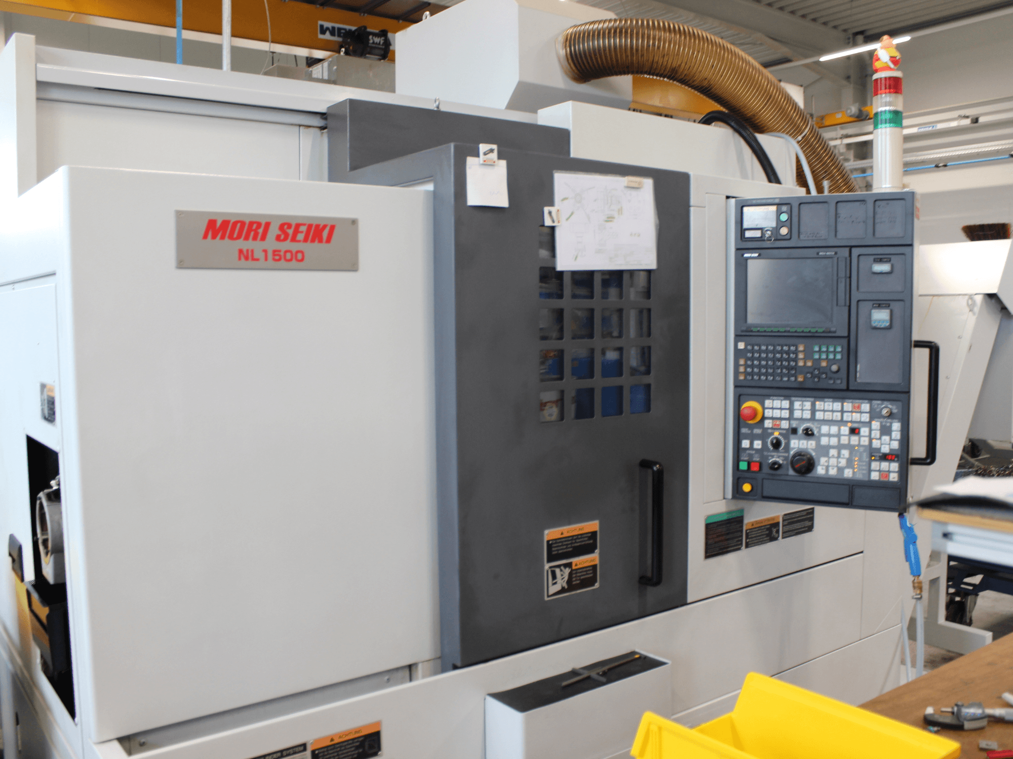 Centro de torneado y fresado MORI SEIKI NL 1500 SY en vista lateral, en el que se aprecian el panel de control, las etiquetas de seguridad operativa y el sistema de refrigeración.