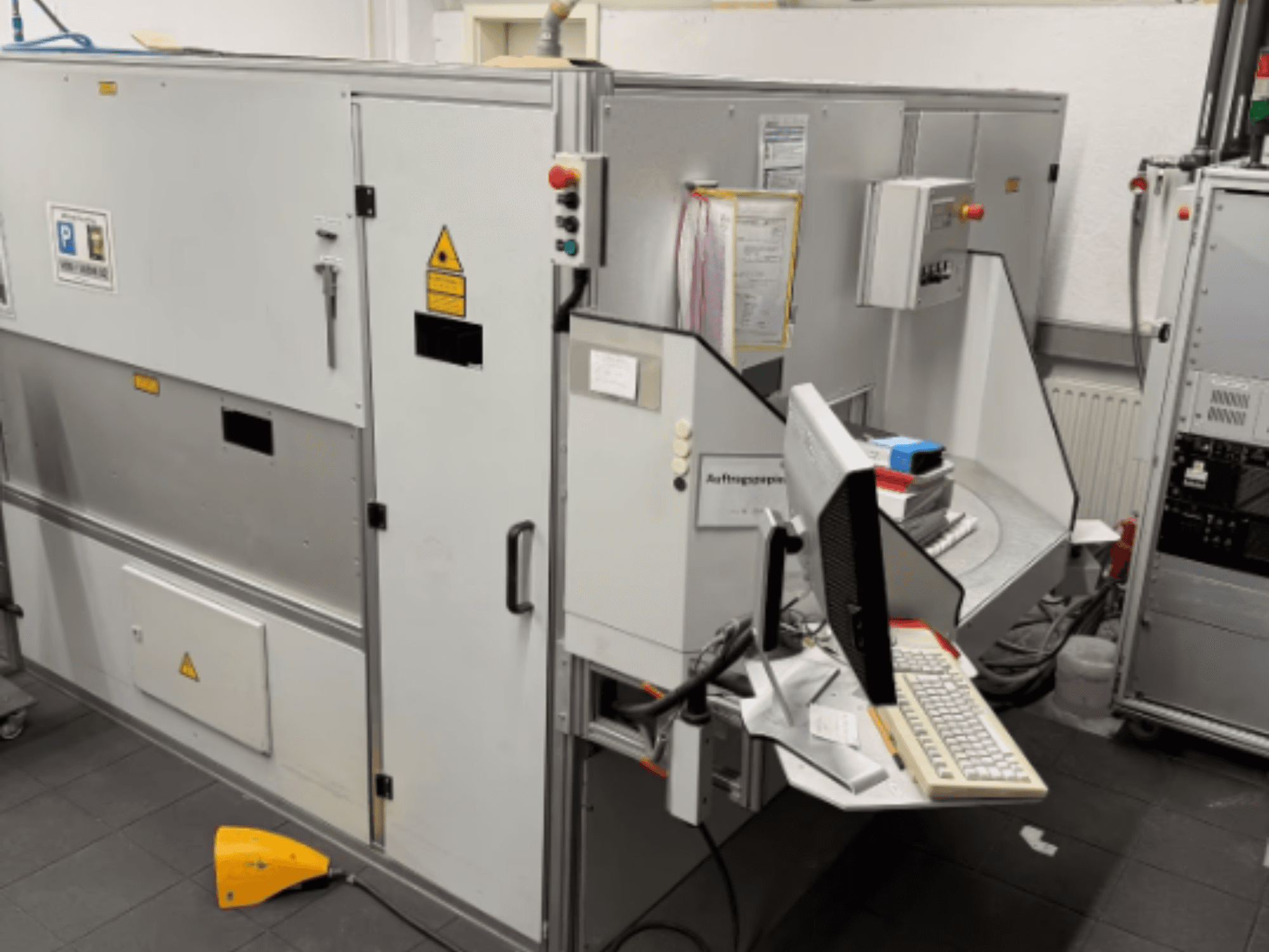 Máquina industrial con un terminal informático, vista lateral; incluye botones de control y señales de advertencia de seguridad.