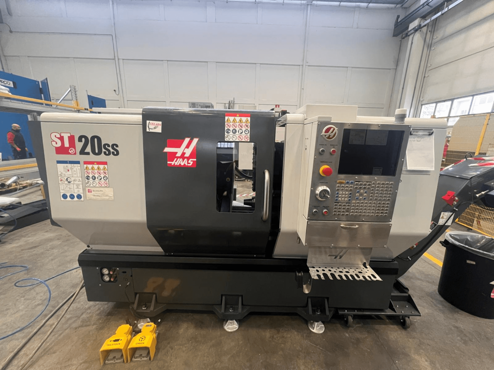 Torno CNC Haas ST 20SS en vista lateral, con panel de control, etiquetas de seguridad y pedales en el suelo.
