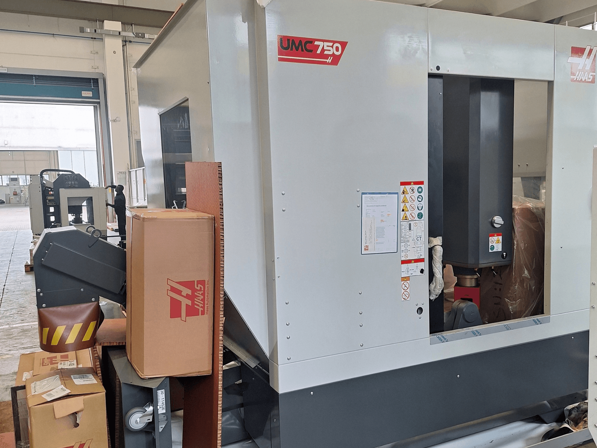 Centro de mecanizado universal HAAS UMC-750, vista lateral, en la que se aprecian el panel de control y el compartimento para herramientas, con etiquetas de advertencia y embalaje.