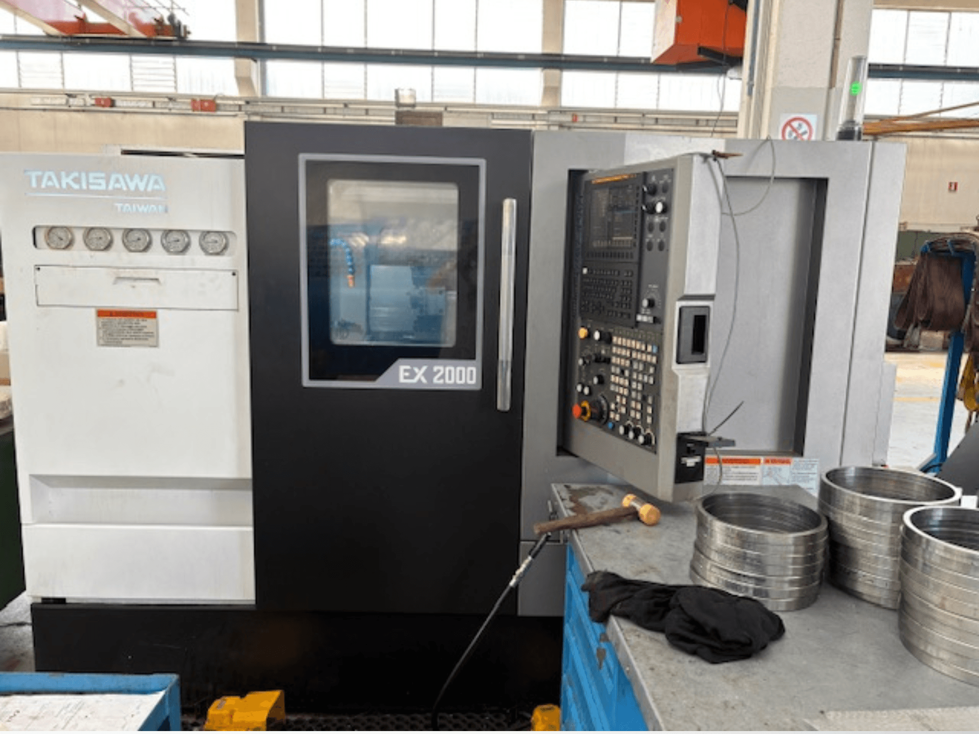 Máquina CNC EX 2000 de Takisawa, vista frontal, con panel de control, calibres y piezas metálicas sobre una mesa de taller.