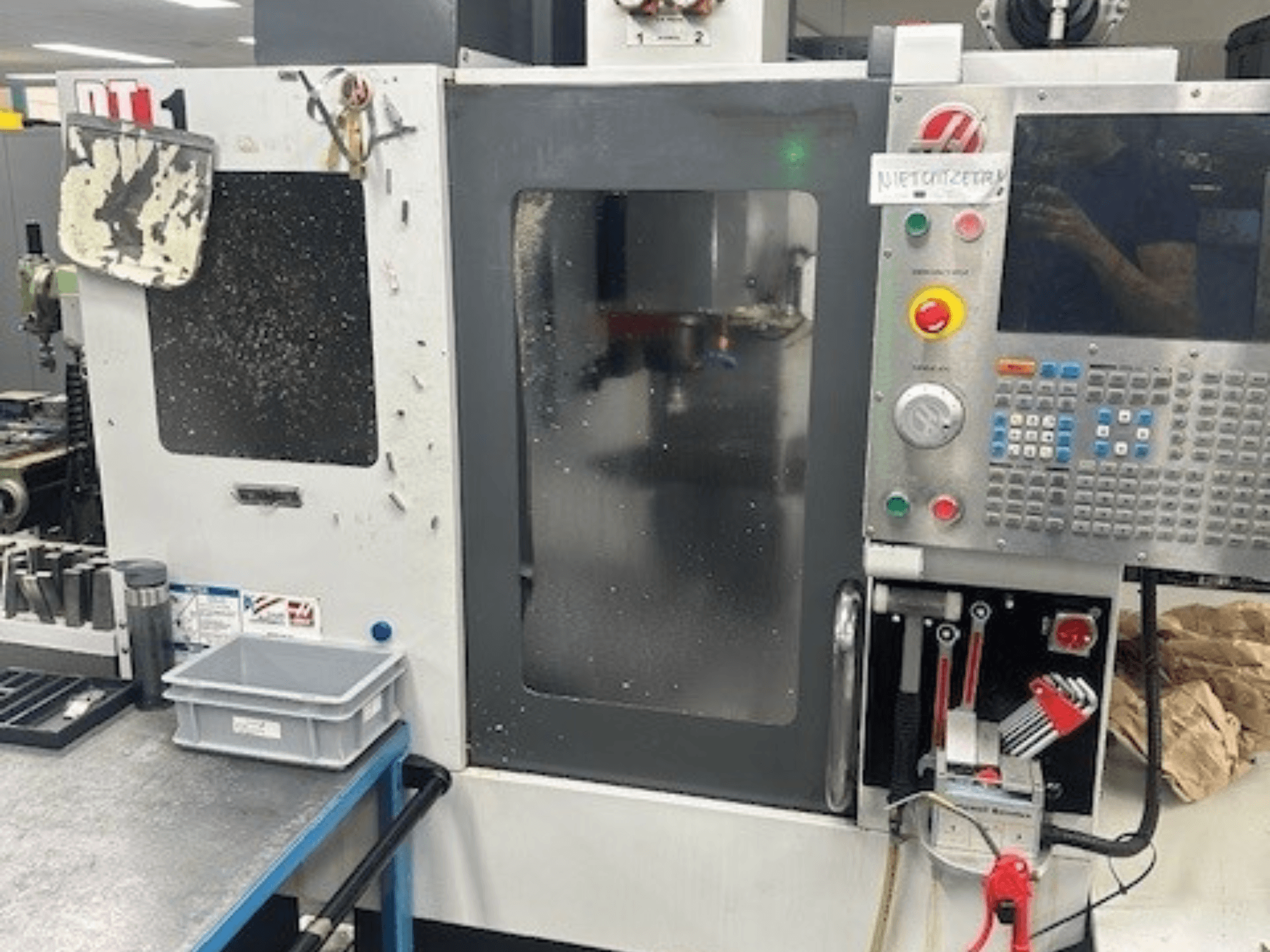 Centro de mecanizado vertical Haas DT-1, vista frontal con panel de control, suciedad visible y bandeja de herramientas en el banco de trabajo.