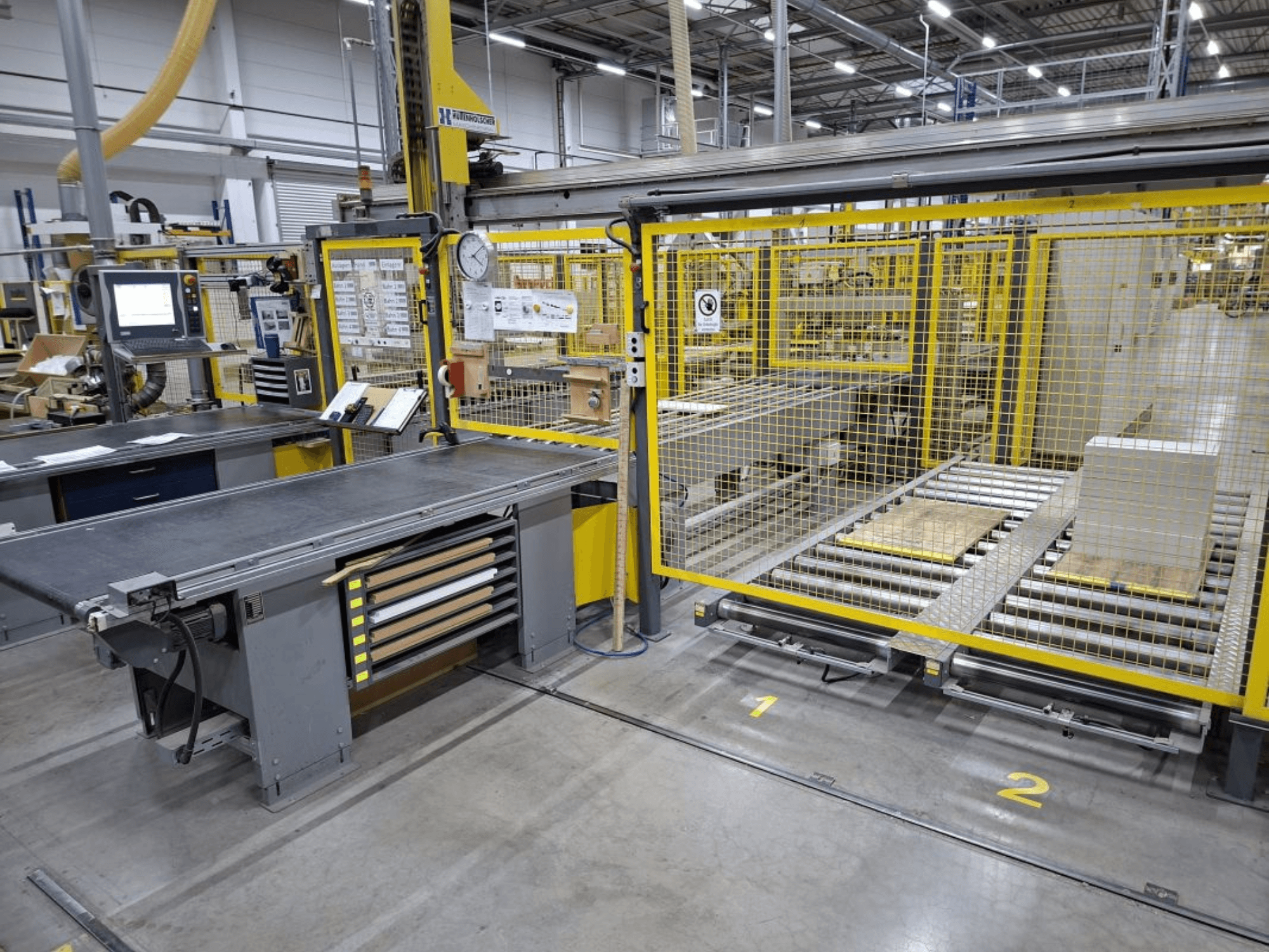 Espacio de trabajo industrial con un sistema transportador y un panel de control; vista lateral que muestra la maquinaria y las vallas de seguridad.