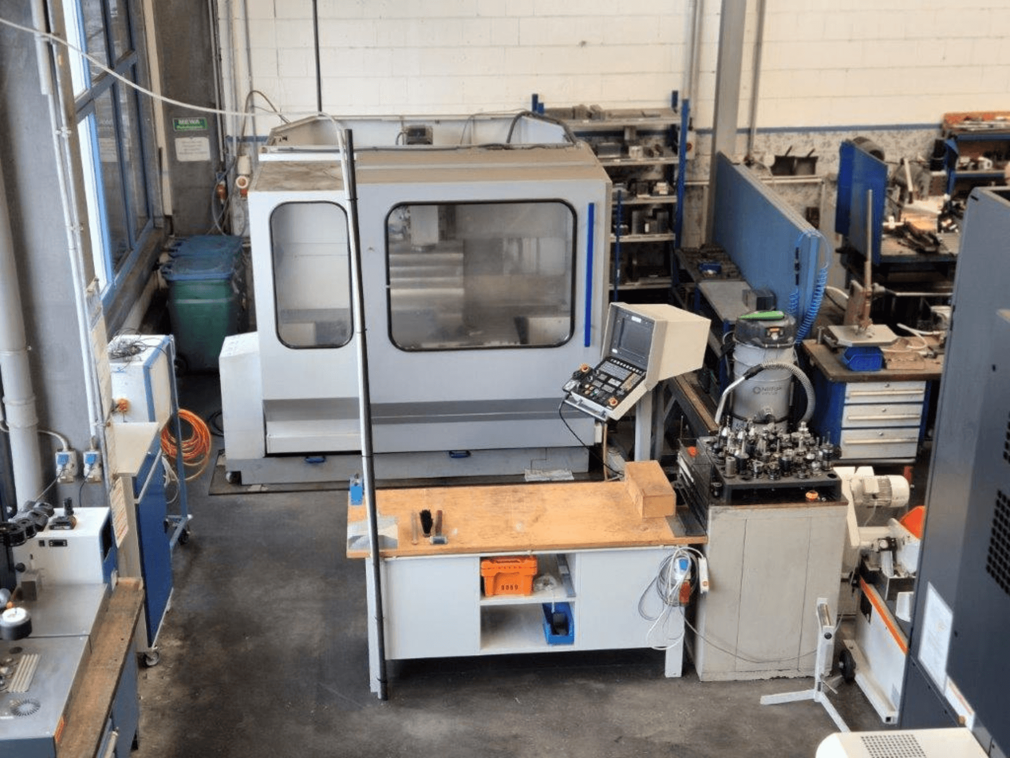 Fresadora CNC en un taller, vista frontal, con panel de control, superficie de trabajo y herramientas visibles al fondo.