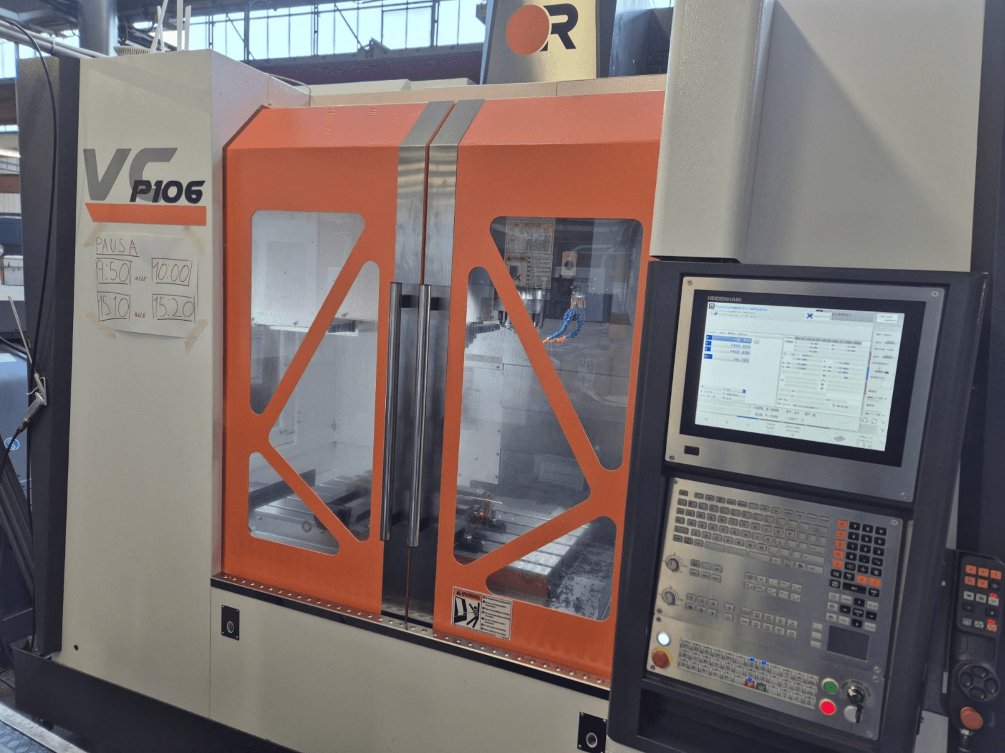 Texto alternativo: Máquina CNC VF P106 naranja y gris, vista frontal, mostrando un panel de control y los tiempos de operación programados en papel.