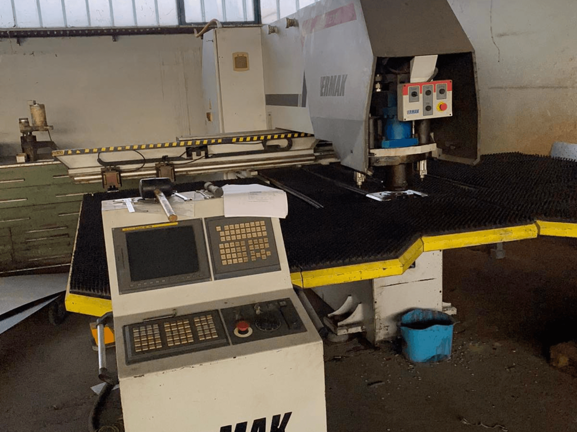 Máquina CNC Ermak, vista frontal, con panel de control, cabezal de corte sobre una alfombrilla de goma y herramientas cercanas en un entorno industrial.