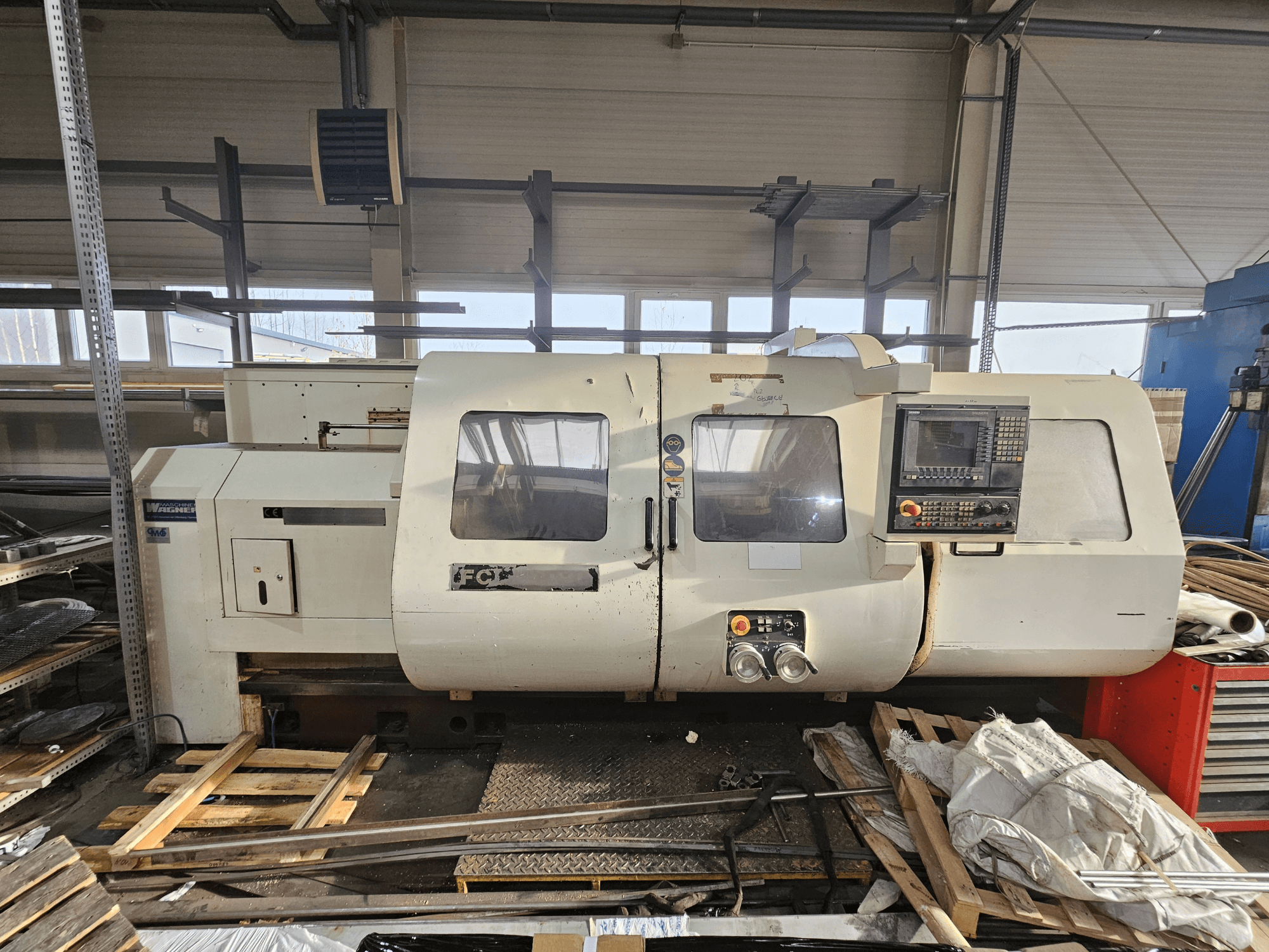 Torno CNC Wabeco, vista lateral, con panel de control, ventanas y área de trabajo; maquinaria sobre palés de madera en un taller.