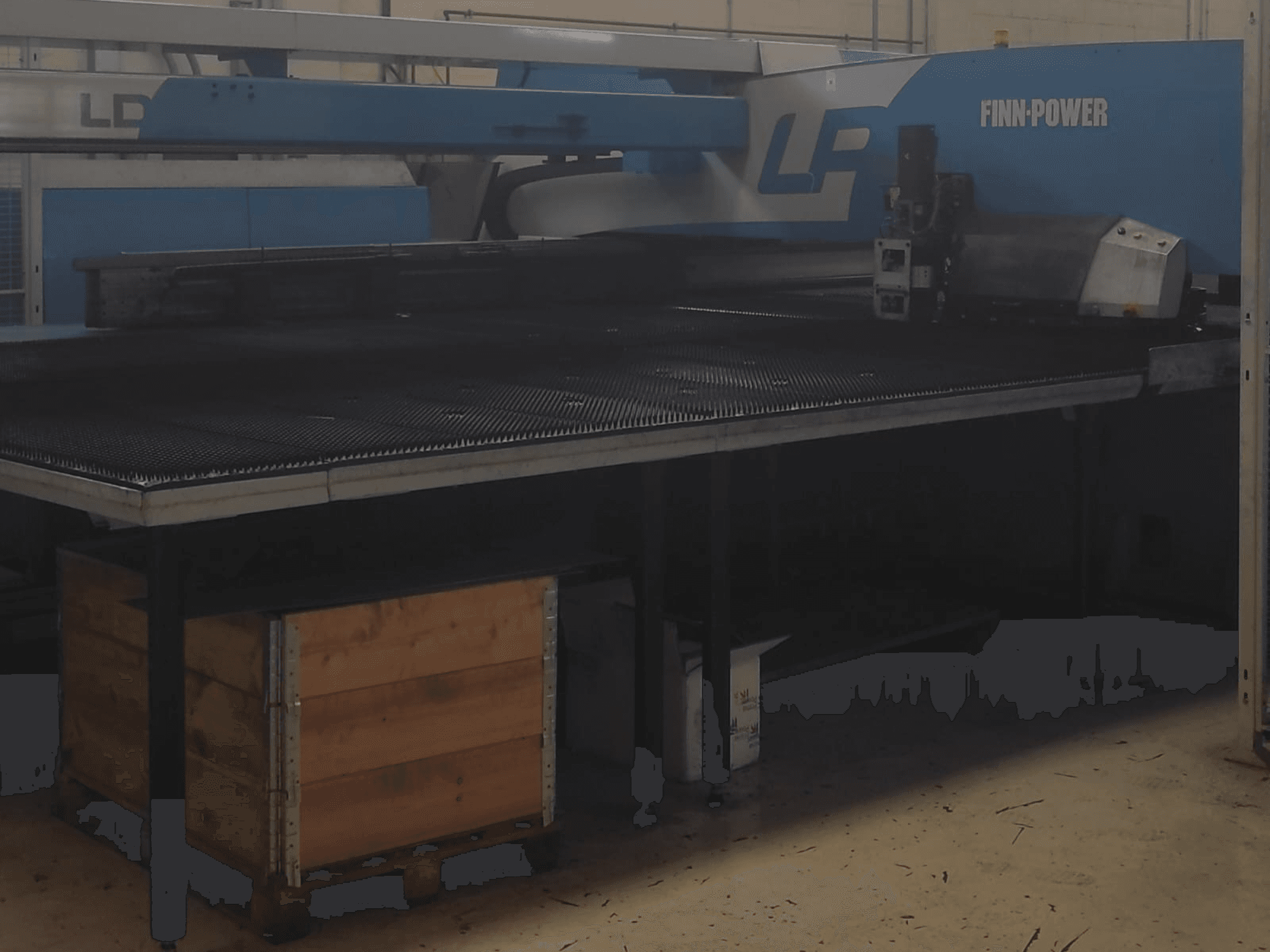 Máquina de corte Finn-Power LP en una fábrica, vista frontal con una mesa metálica texturizada y una caja de madera debajo.