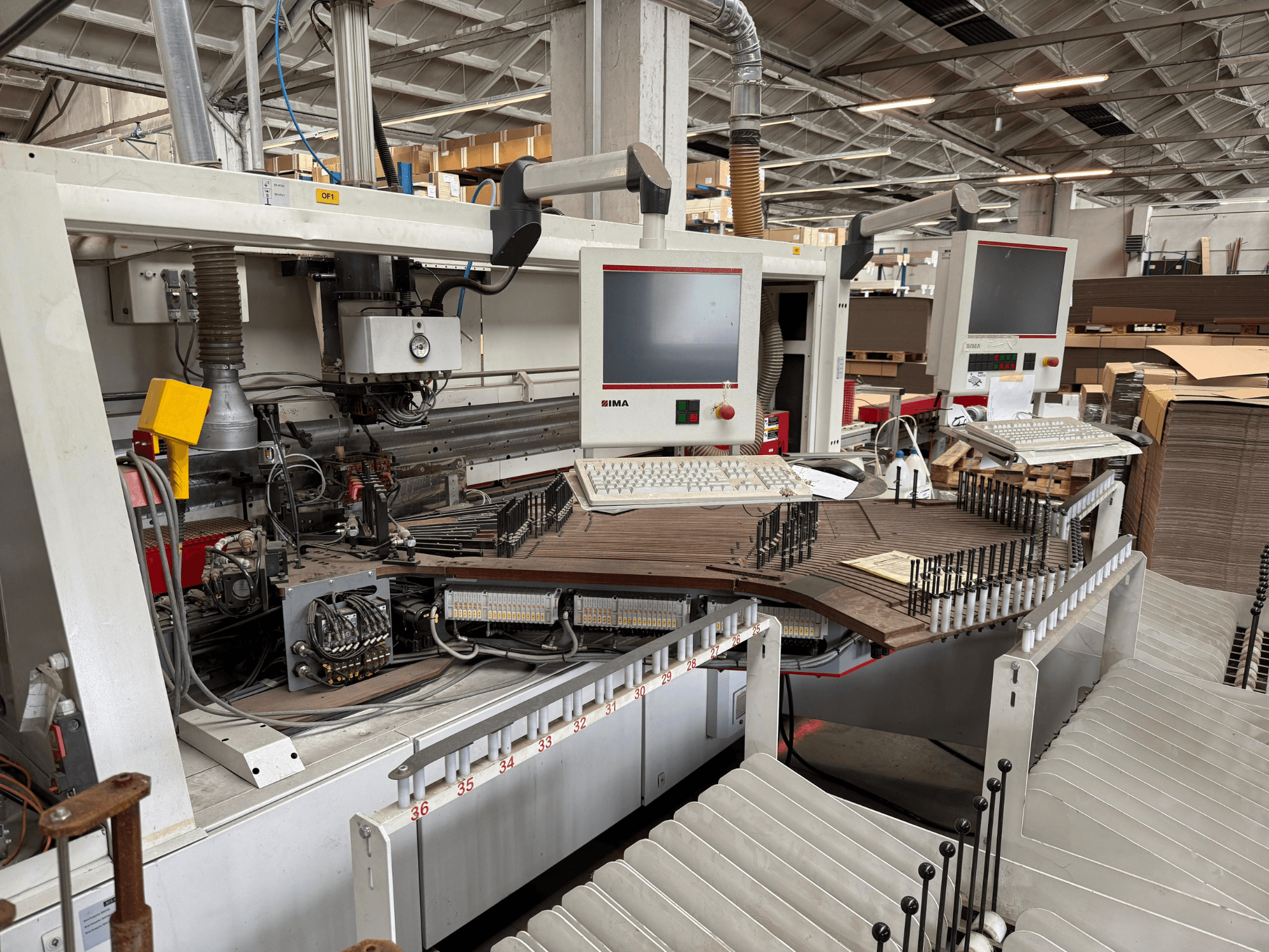 Máquina de precisión IMA para trabajar la madera con dos monitores y paneles de control, vista frontal que muestra el transportador y la zona de herramientas.