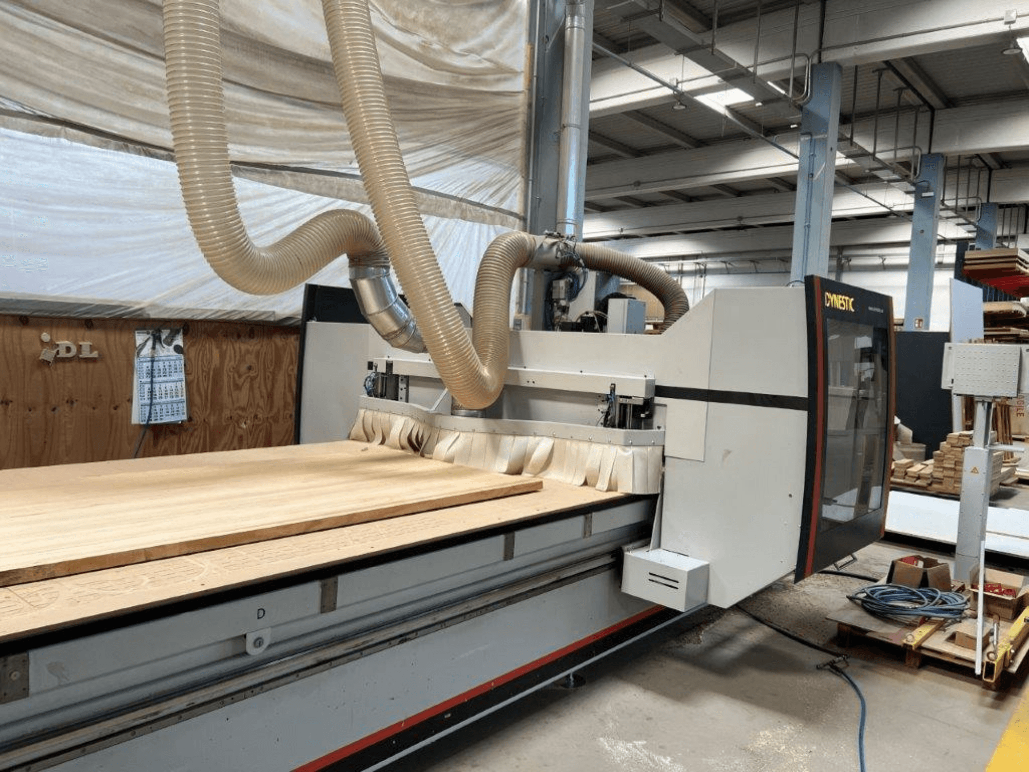 Máquina CNC de Dynestic, vista frontal, con una gran pieza de trabajo de madera y manguera de extracción de polvo en un entorno de taller.