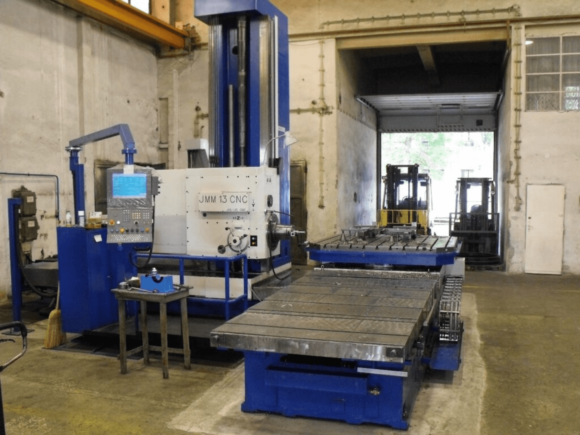 Fresadora CNC JMM 13 en un taller, vista frontal, cuenta con un panel de control, una gran mesa de trabajo y un fondo industrial.