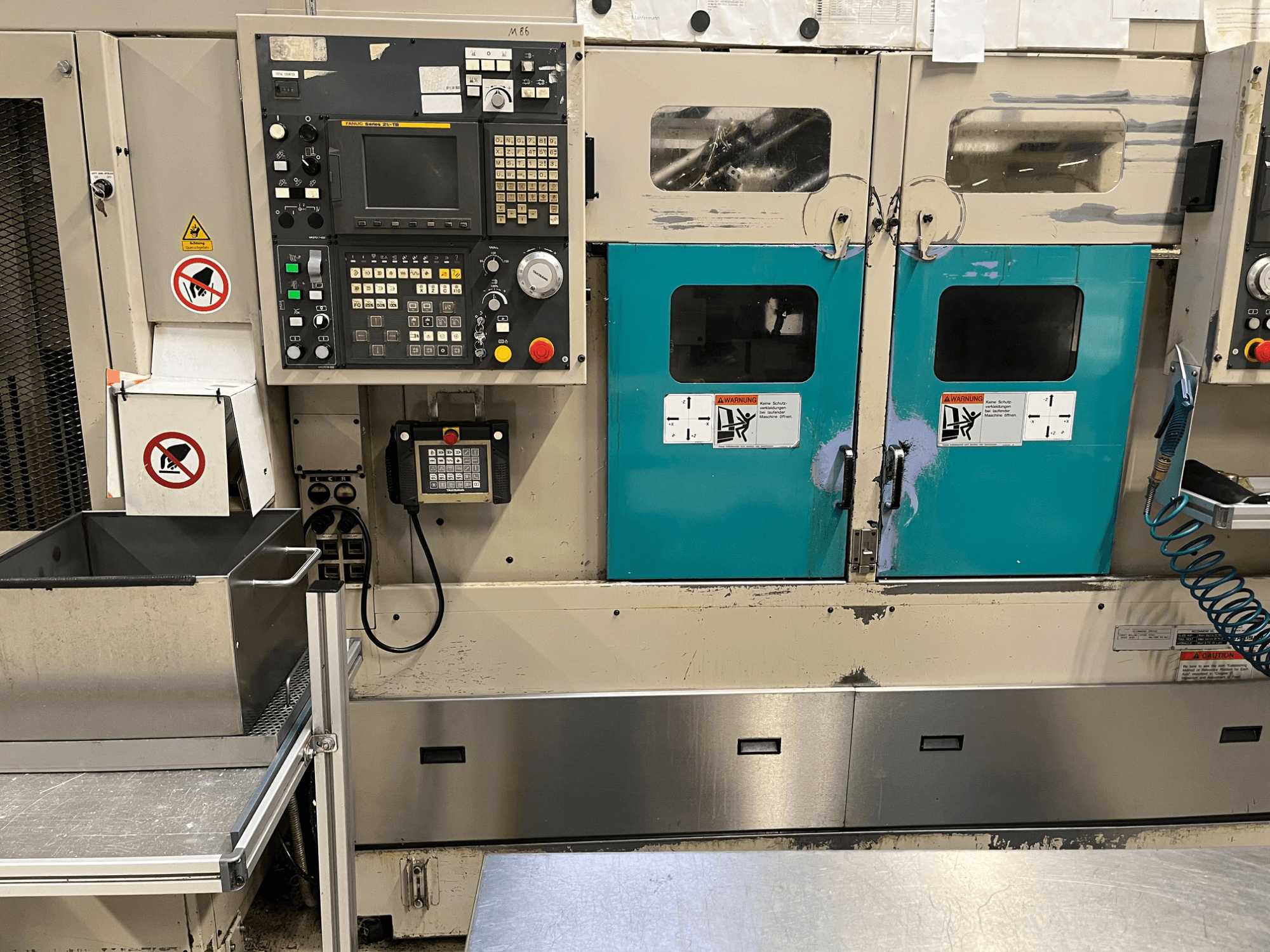 Panel de control de una máquina de fabricación con puertas de color verde azulado, advertencias de seguridad y un teclado; vista frontal, acabado en acero inoxidable.