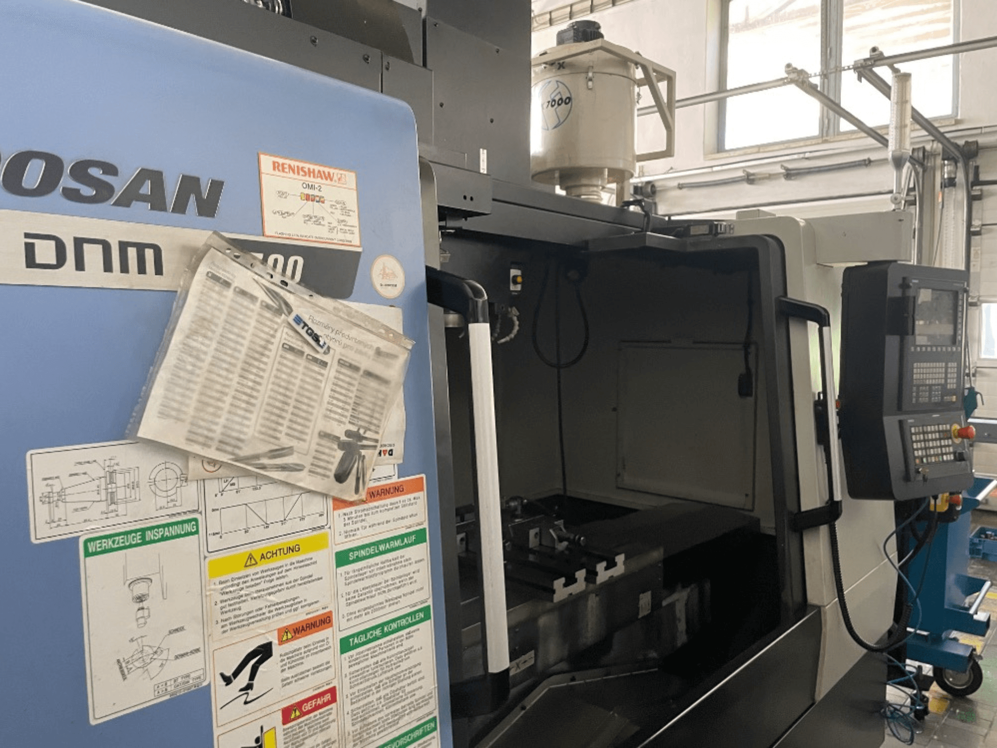 Centro de mecanizado vertical DOOSAN DNM 5700, vista frontal, muestra panel de control, etiquetas de seguridad y detalles de la máquina.