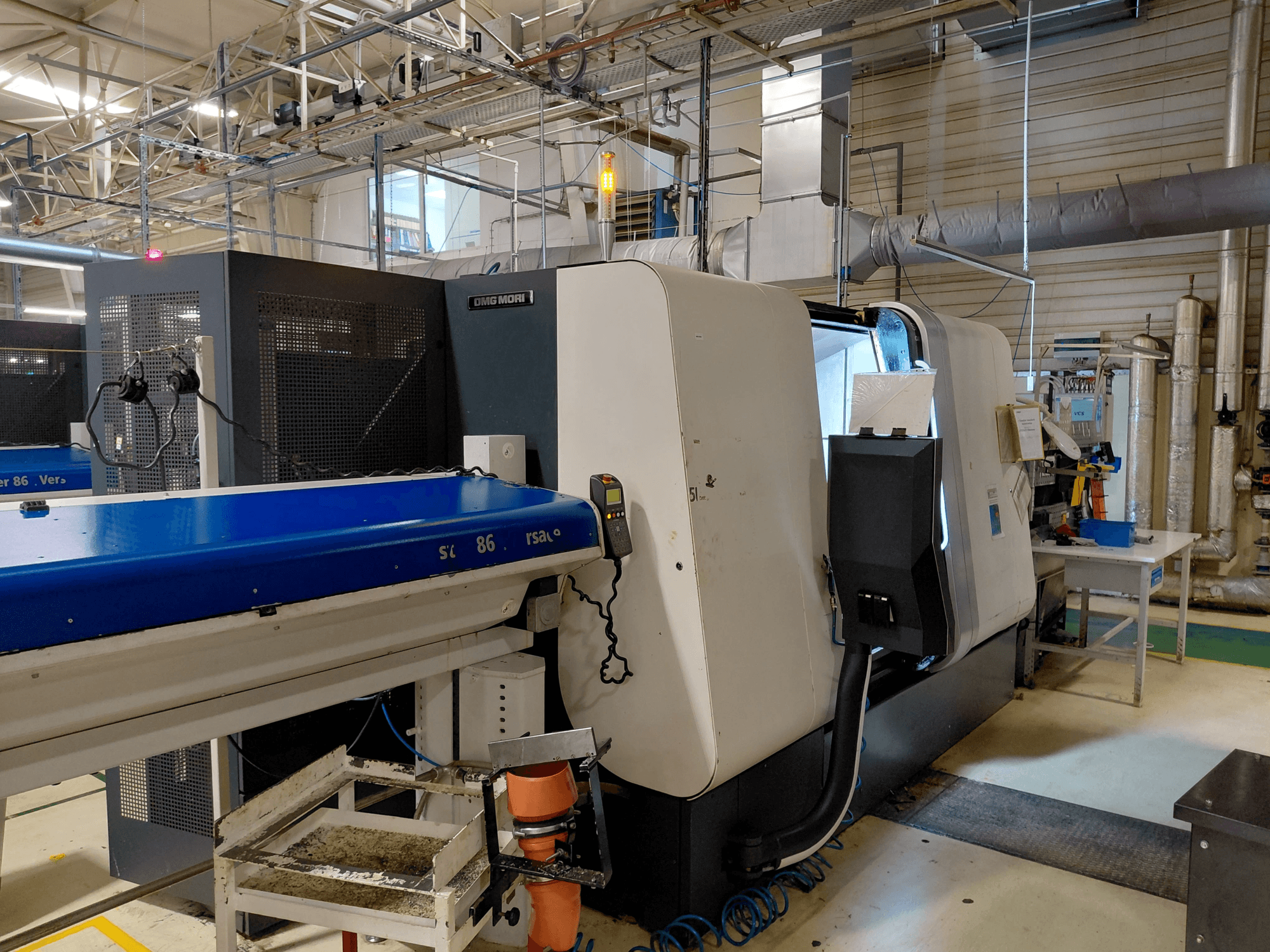 Máquina de torneado multihusillo DMG MORI Sprint 65 linear, vista lateral con panel de control y cinta transportadora visibles en un taller.