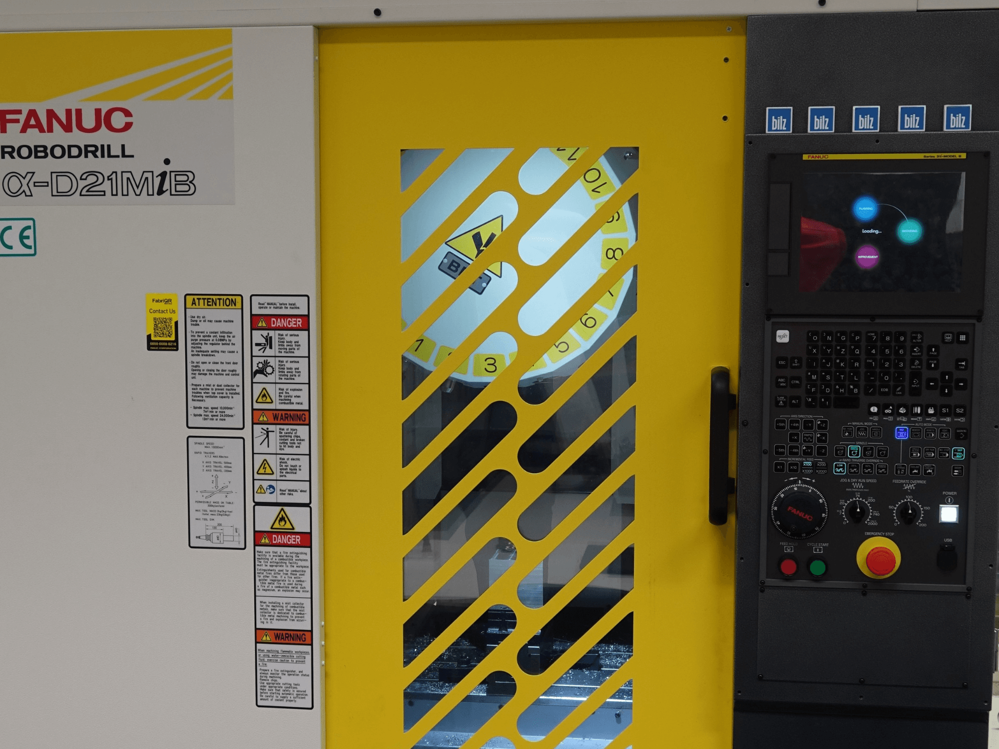 Vista frontal del centro de mecanizado vertical FANUC α-D21MiB, en la que se aprecian las advertencias de seguridad, el panel de control y la puerta de seguridad amarilla.