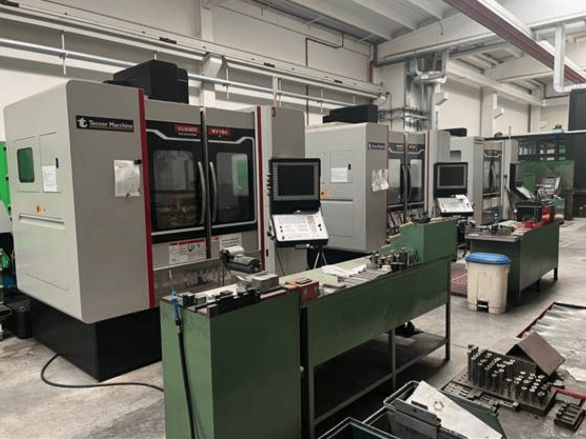 Cuatro máquinas CNC de Tecnoer Marchine en un taller, vista frontal, con herramientas y piezas en bancos de trabajo verdes.