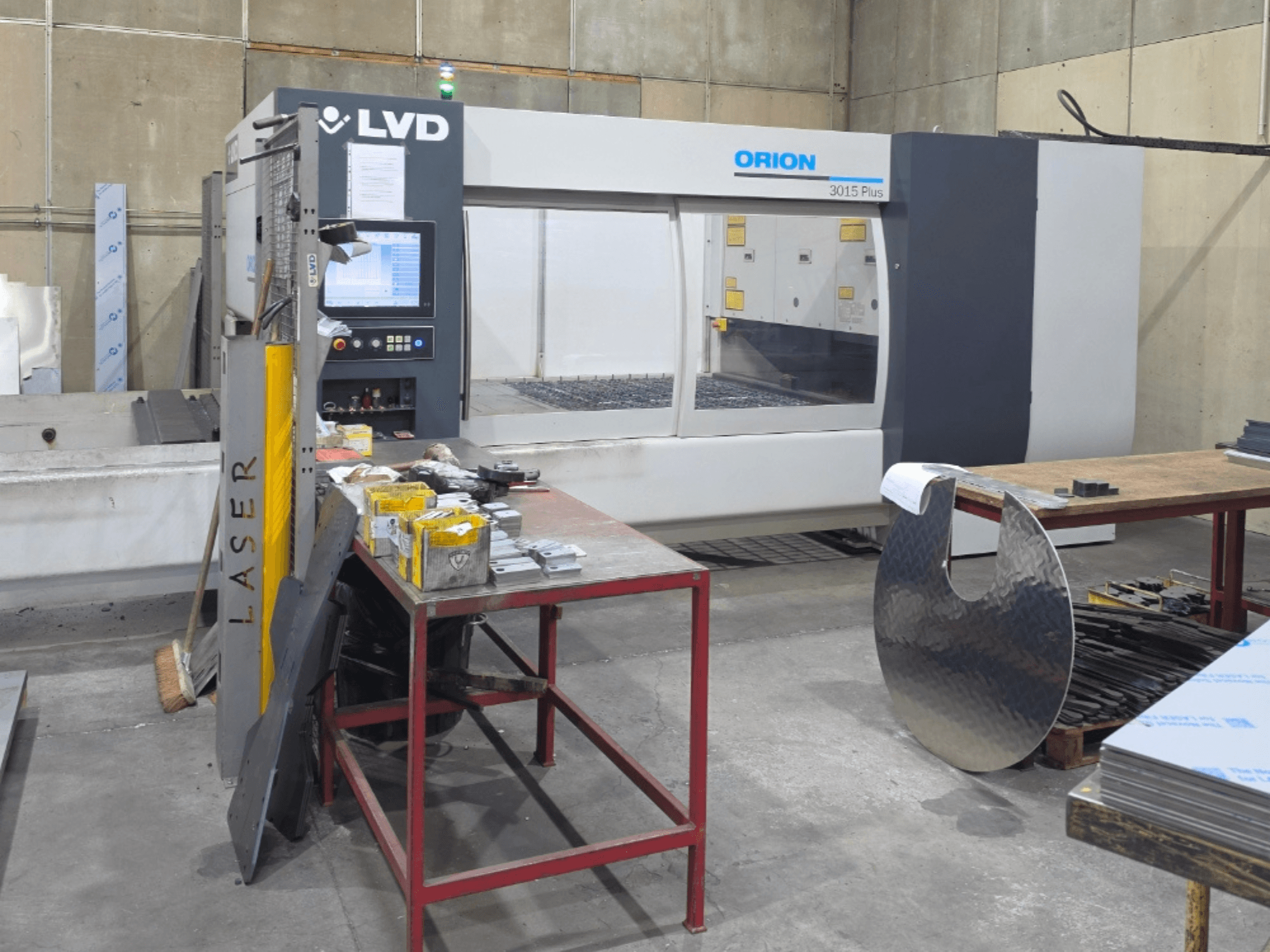 Máquina de corte por láser de CO₂ LVD Orion 3015 Plus, vista frontal con el panel de control; rodeada de piezas metálicas sobre una mesa de trabajo.