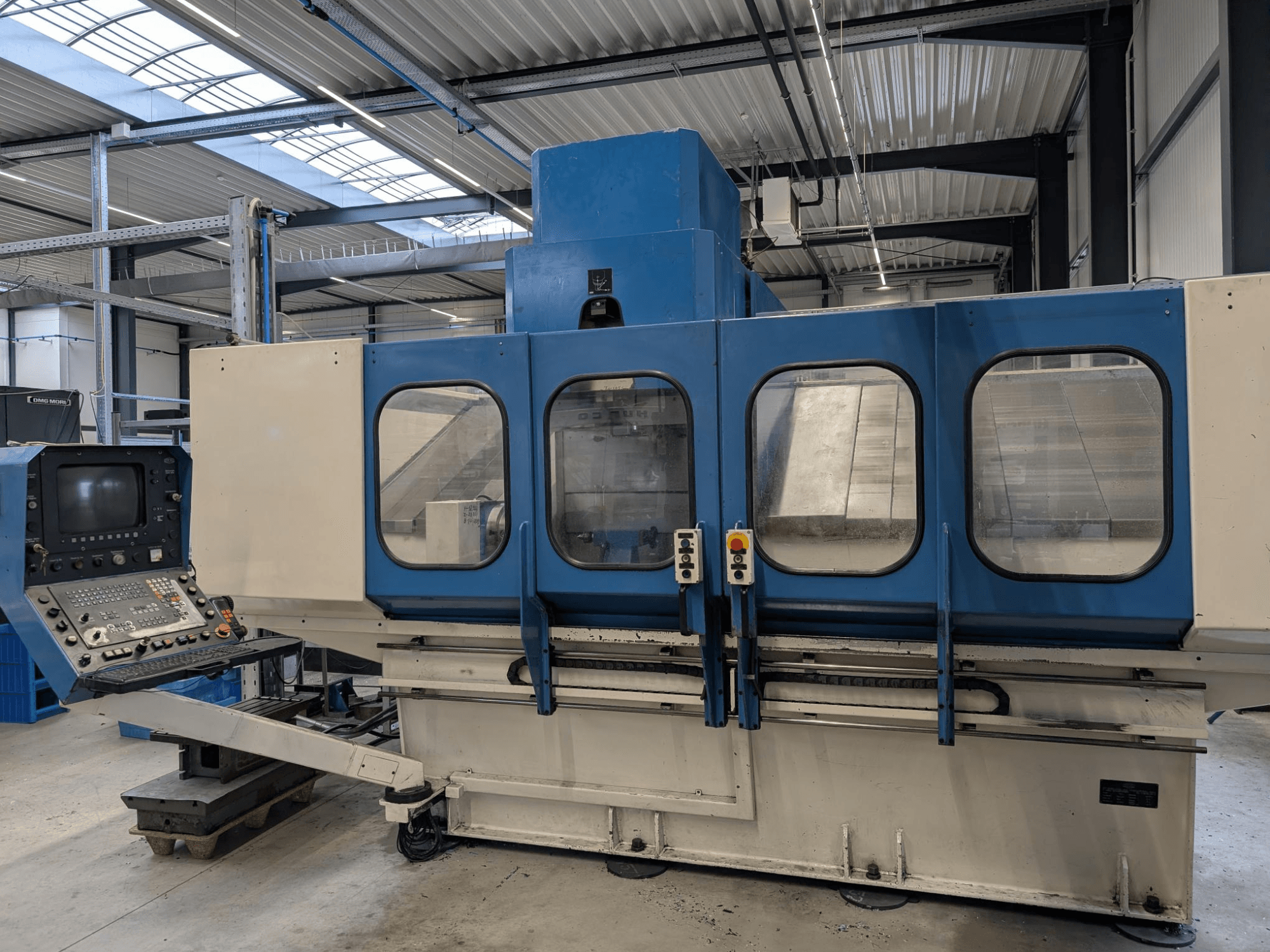 Máquina CNC industrial azul con panel de control, vista frontal. Grandes ventanas de cristal, componentes de maquinaria y botones visibles.