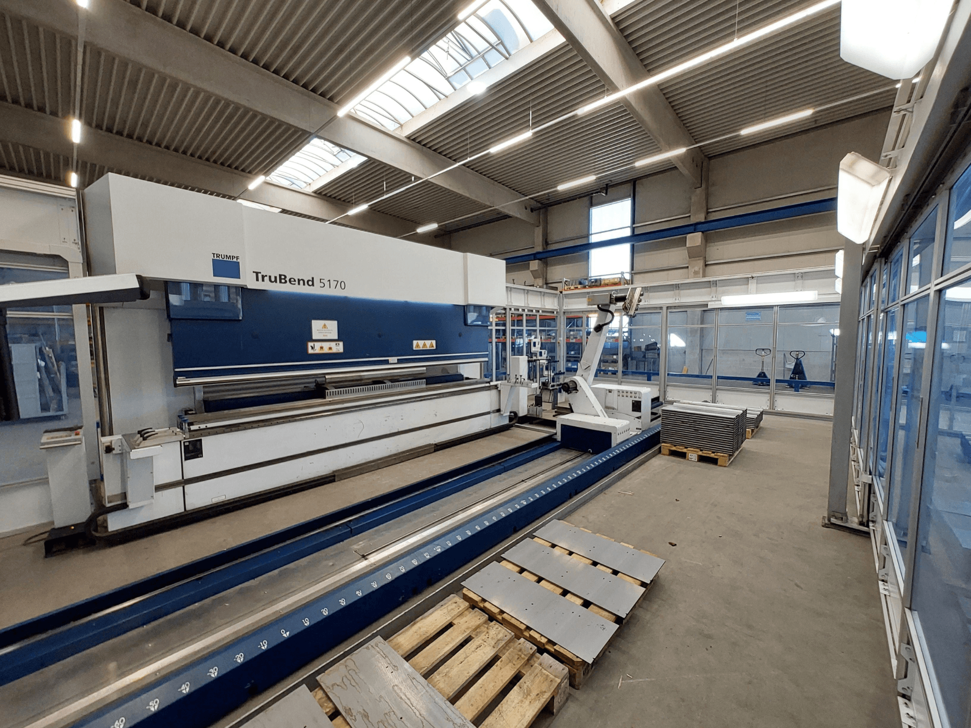 Plegadora TRUMPF TruBend 5170 en un taller, vista lateral, mostrando la herramienta de plegado y chapas apiladas en palés.