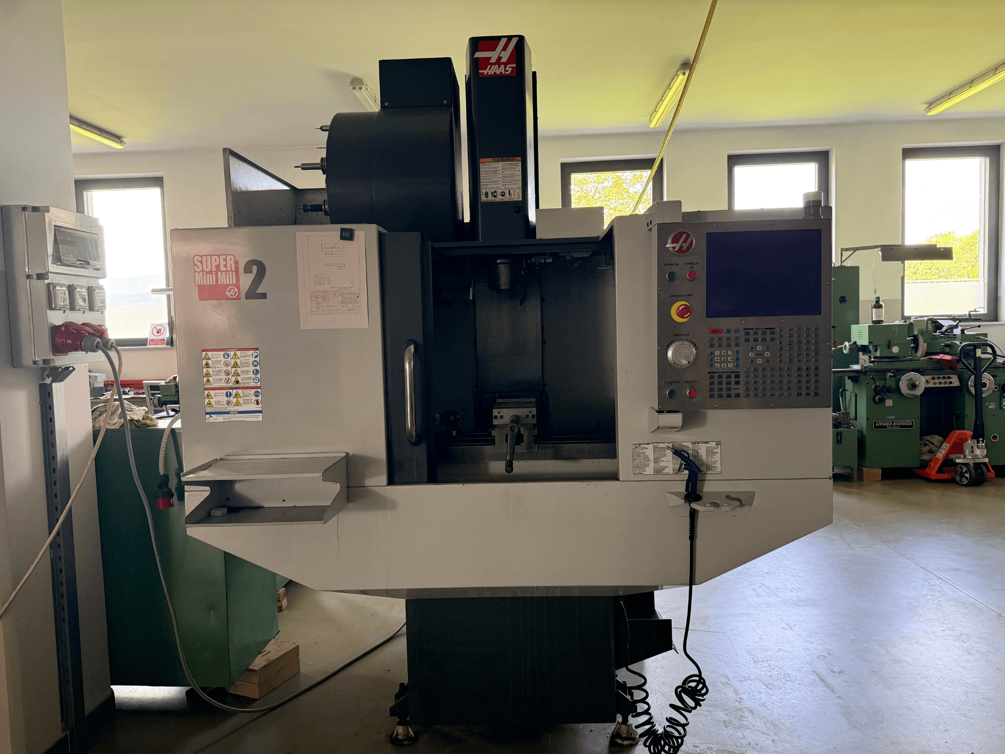 Centro de mecanizado vertical Haas Mini Mill 2 en vista frontal, mostrando el panel de control y la zona de herramientas con etiquetas de advertencia.
