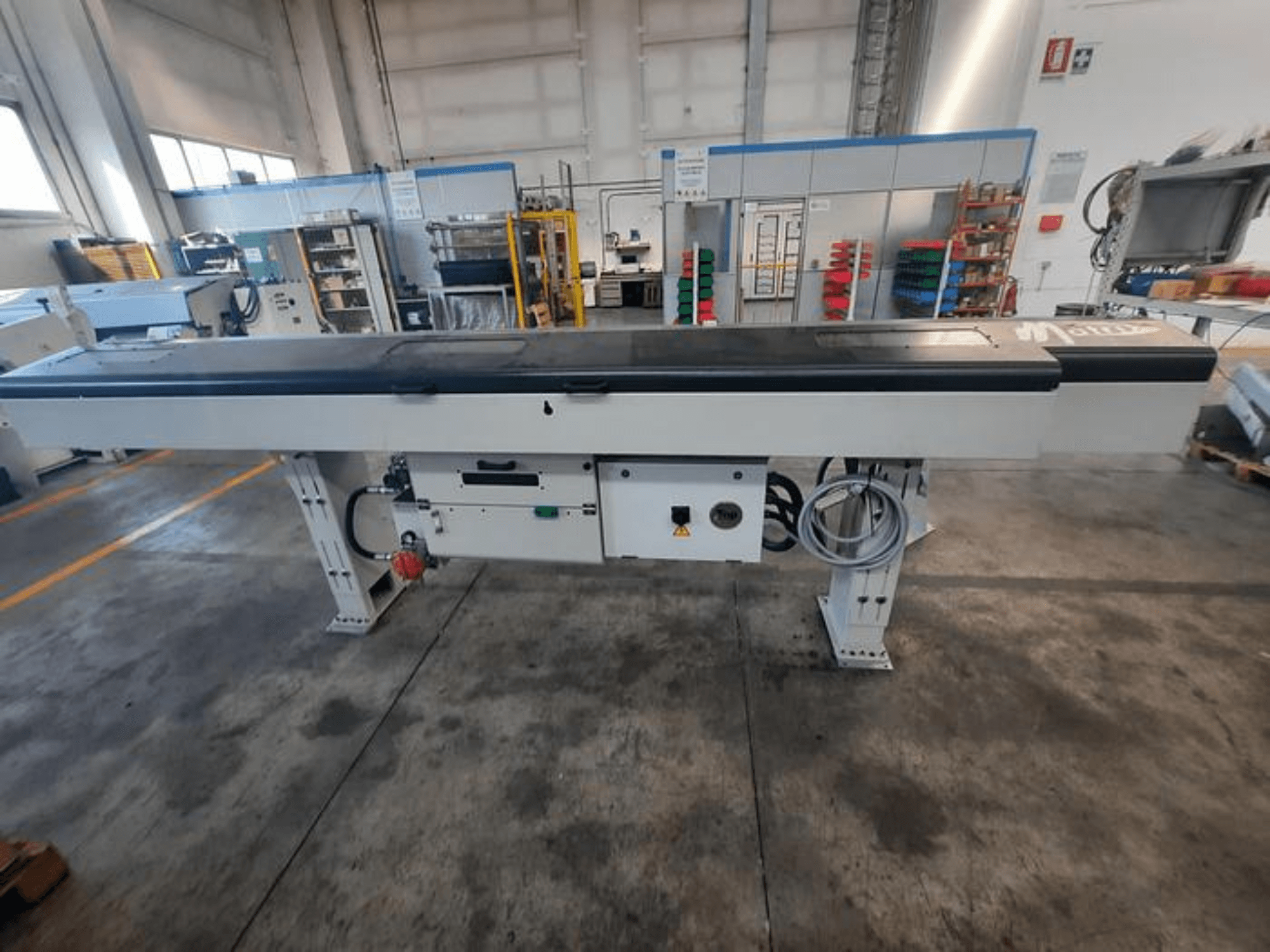 Mesa transportadora metálica con tablero negro liso, vista lateral, con botones de control y cables; logotipo de la marca parcialmente visible.