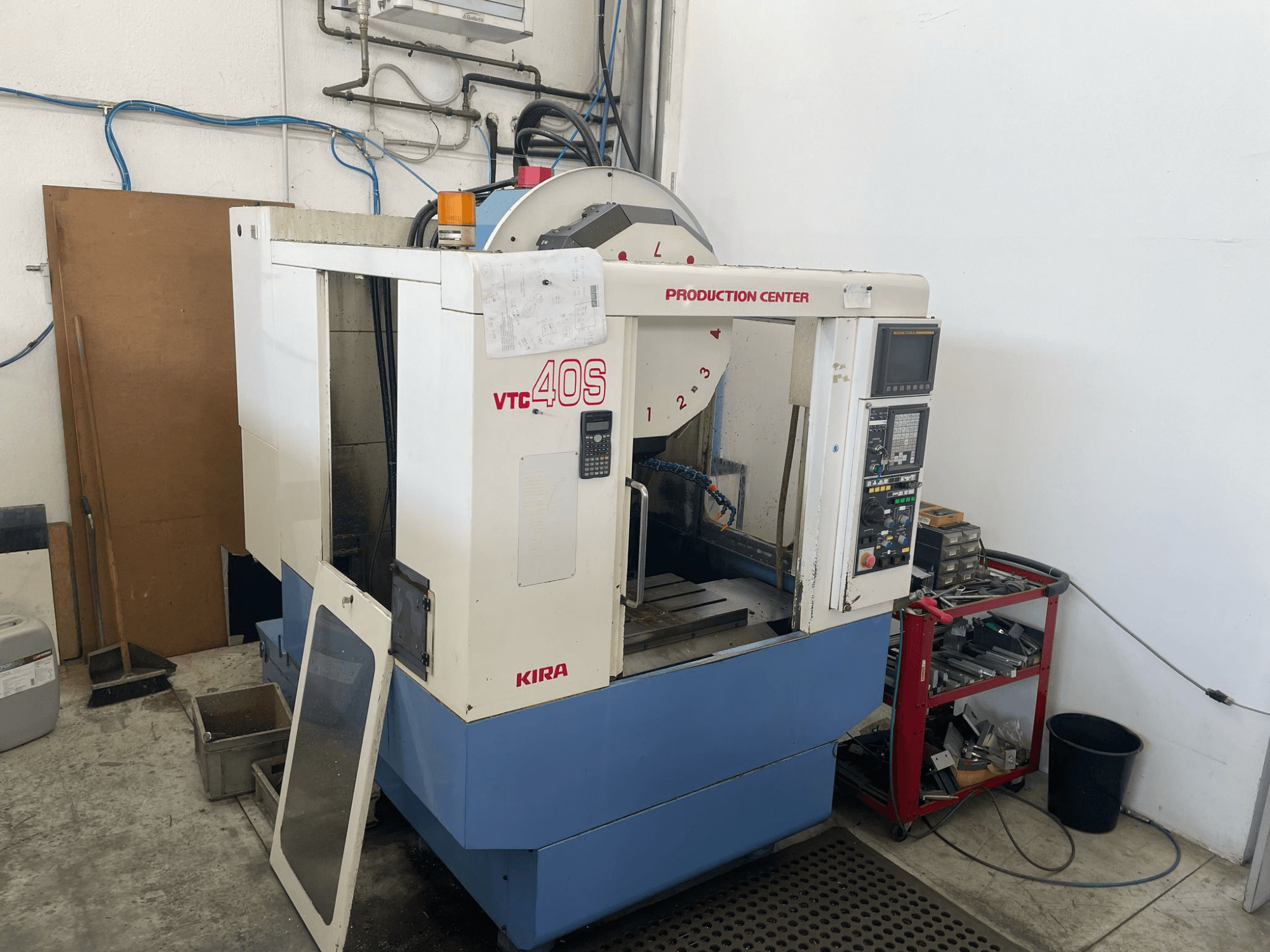 Centro de mecanizado KIRA VTC 40S en vista lateral, con panel de control, puerta abierta y herramientas en un carro de almacenamiento cercano.