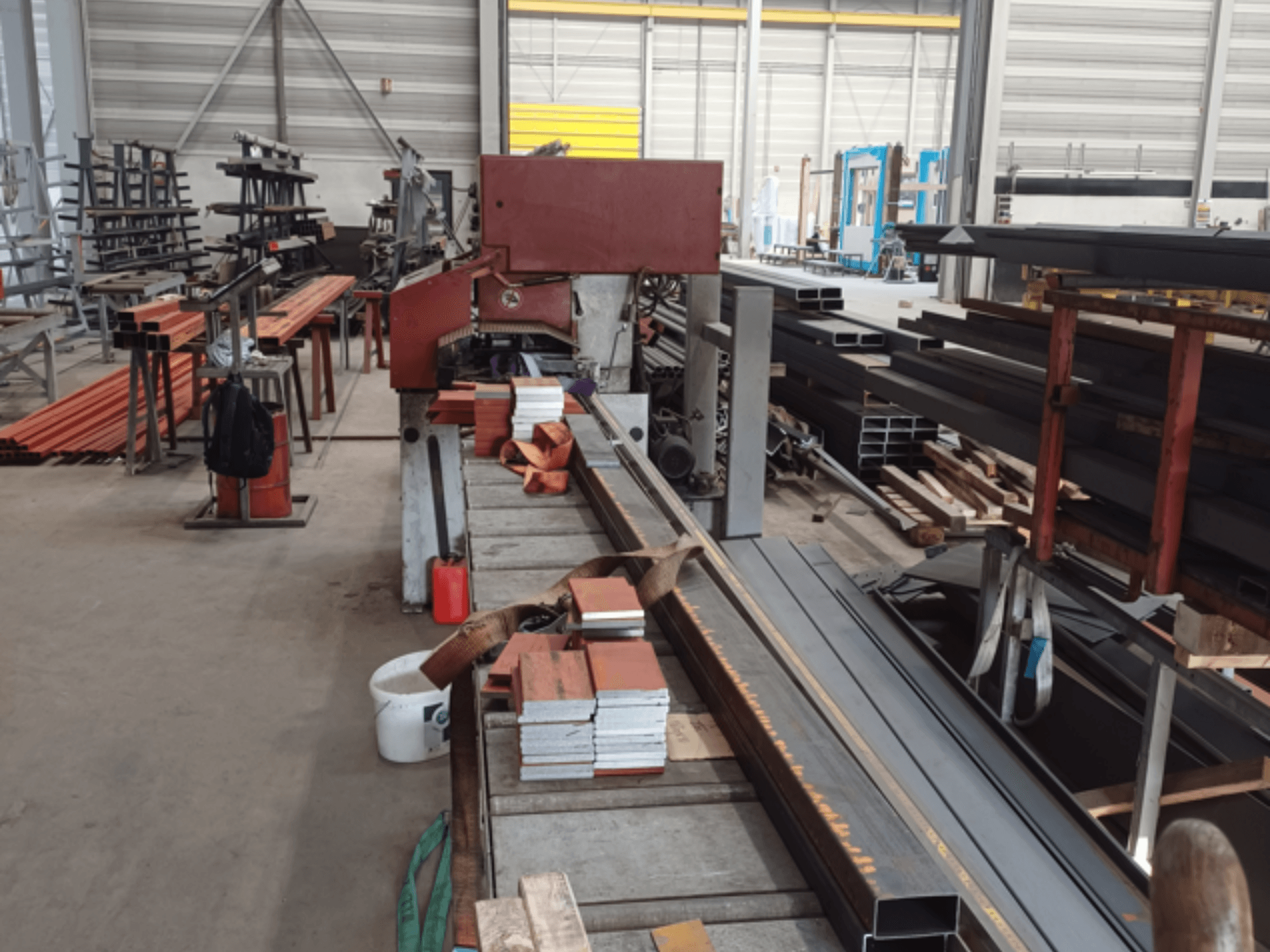 Taller de fabricación de diversos perfiles metálicos, con una sierra roja en primer plano y materiales apilados.