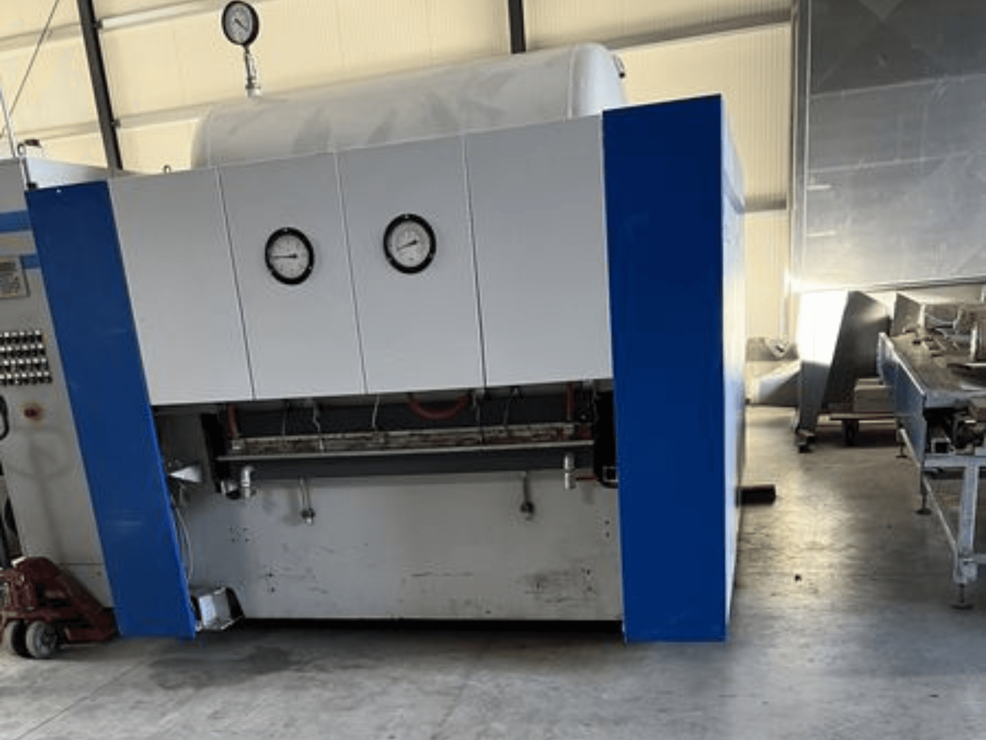 Máquina industrial azul y blanca con dos manómetros, vista frontal, con panel de control y equipos adyacentes.