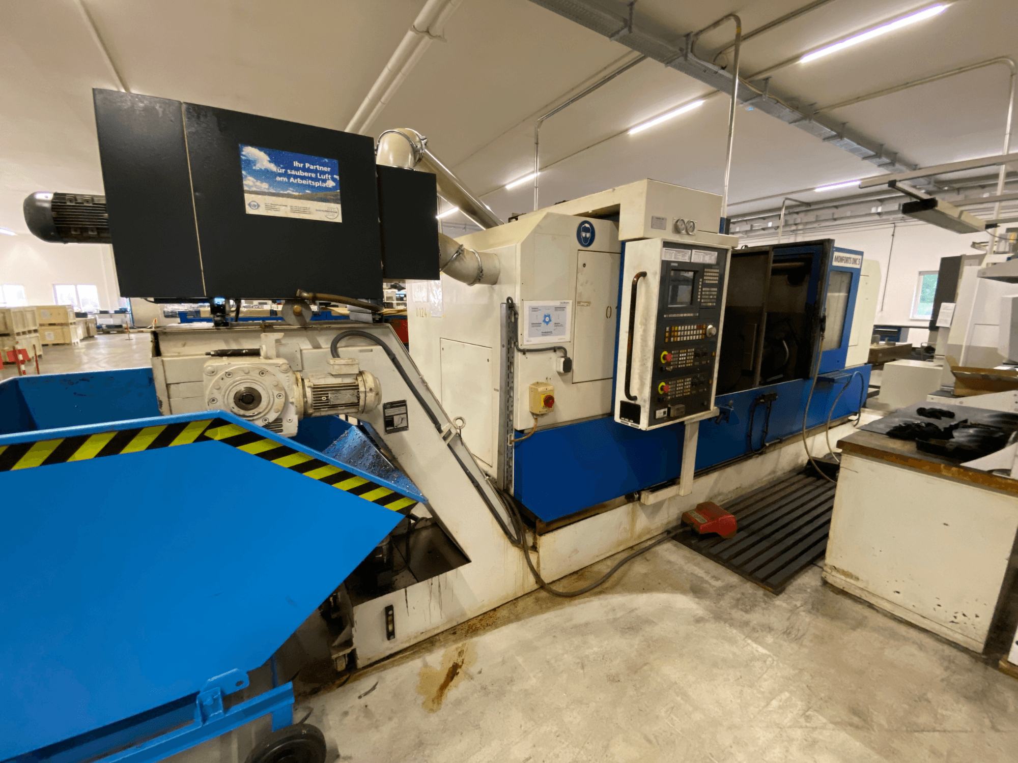 Centro de torneado y fresado Monforts DNC 5, vista lateral en la que se aprecian el panel de control, la bandeja azul para virutas y detalles de la maquinaria en un entorno de taller.