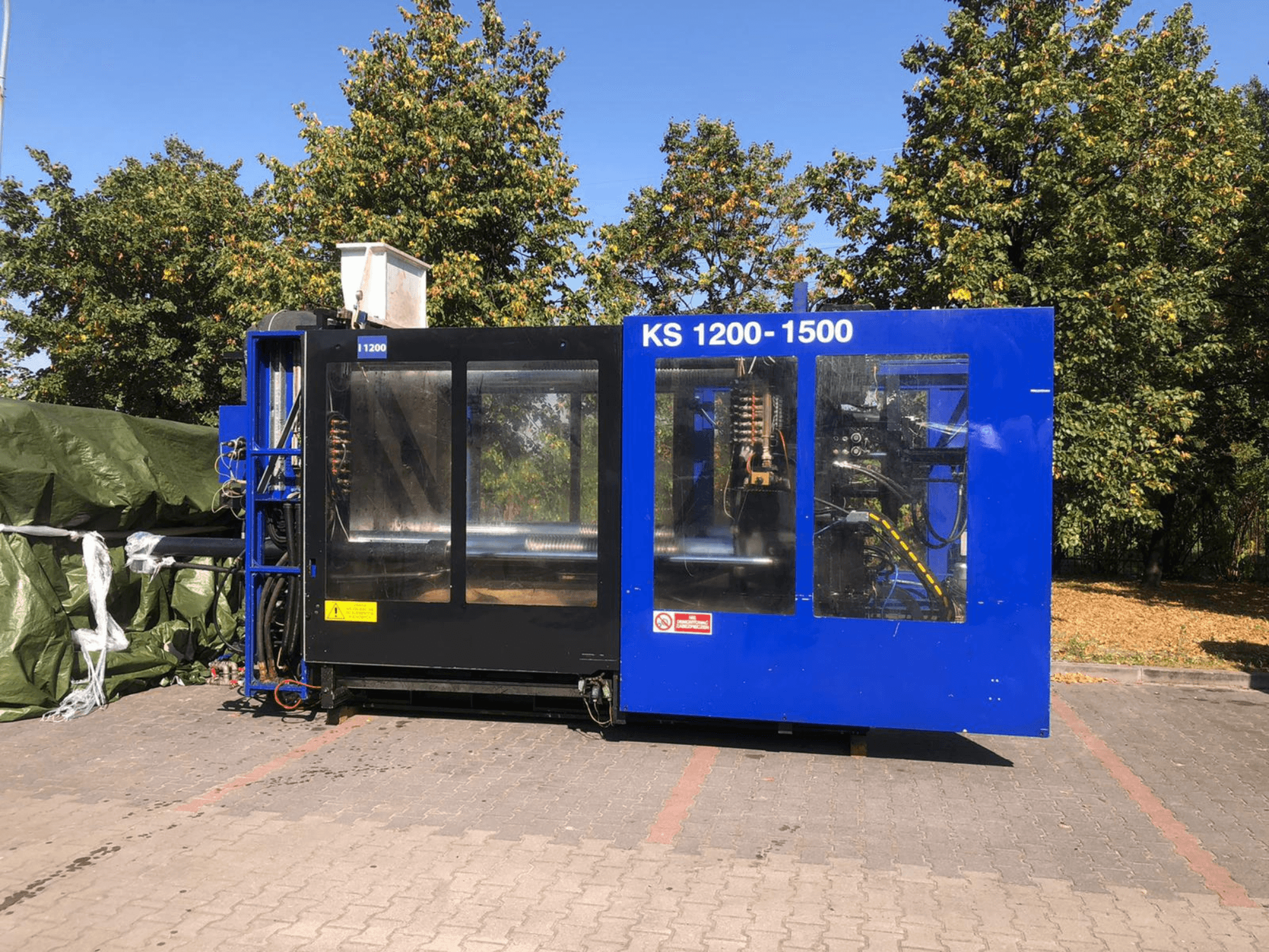 Máquina de Moldeo por Inyección Hidráulica ITALTECH KS 1200 8300 ES, vista lateral, mostrando controles y detalles de la maquinaria en un entorno de parque.