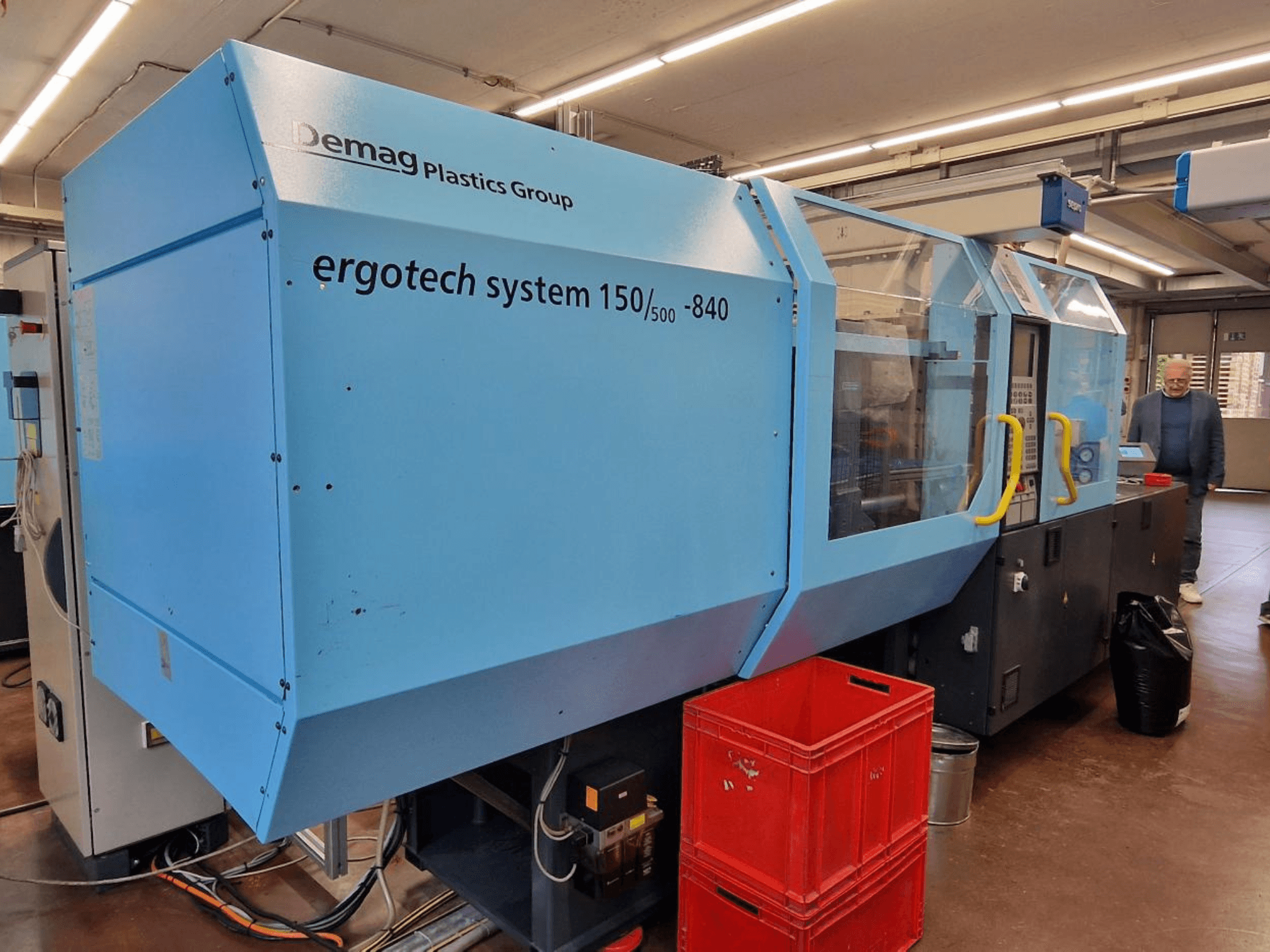 Máquina de moldeo por inyección Demag Ergotech System 150/500-840 en azul, vista lateral, con cajas y operario al fondo.