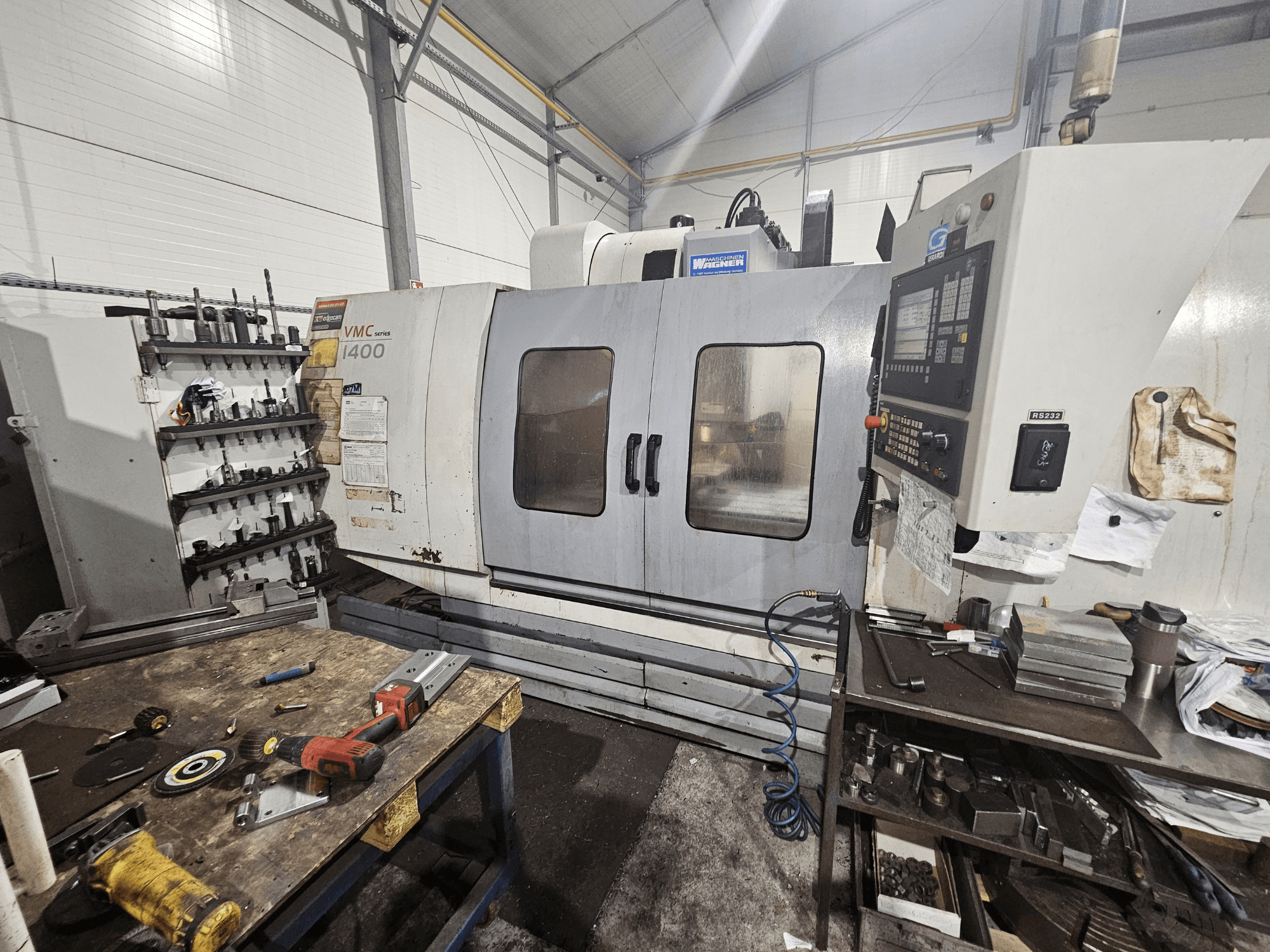 Fresadora CNC VMC 1400 en un taller, vista frontal; herramientas y materiales esparcidos en el banco de trabajo cercano.
