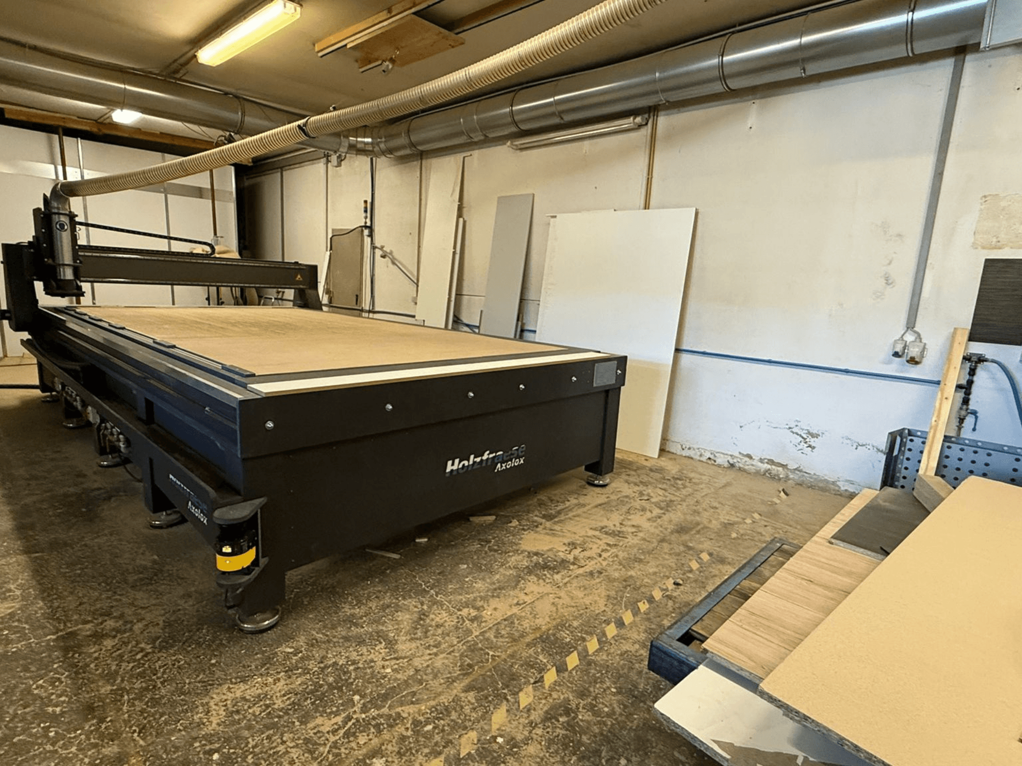Fresadora CNC Holz-Her Axos 5 en el taller, vista frontal mostrando la mesa de trabajo, el sistema de extracción de polvo y los paneles de madera cercanos.
