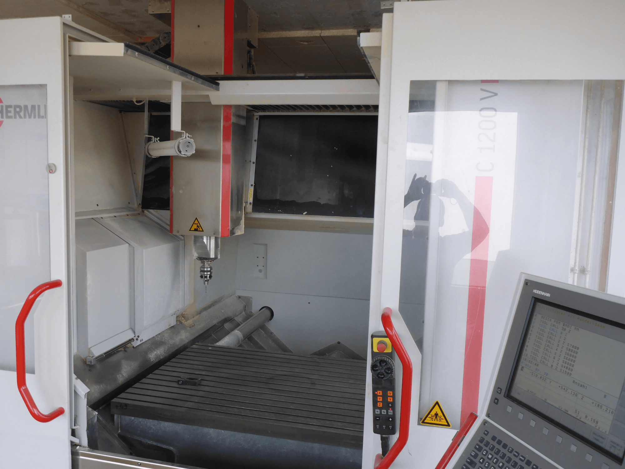 Interior del centro de mecanizado vertical HERMLE C 1200 mostrando el portaherramientas, el panel de control y la mesa de trabajo desde una vista frontal.