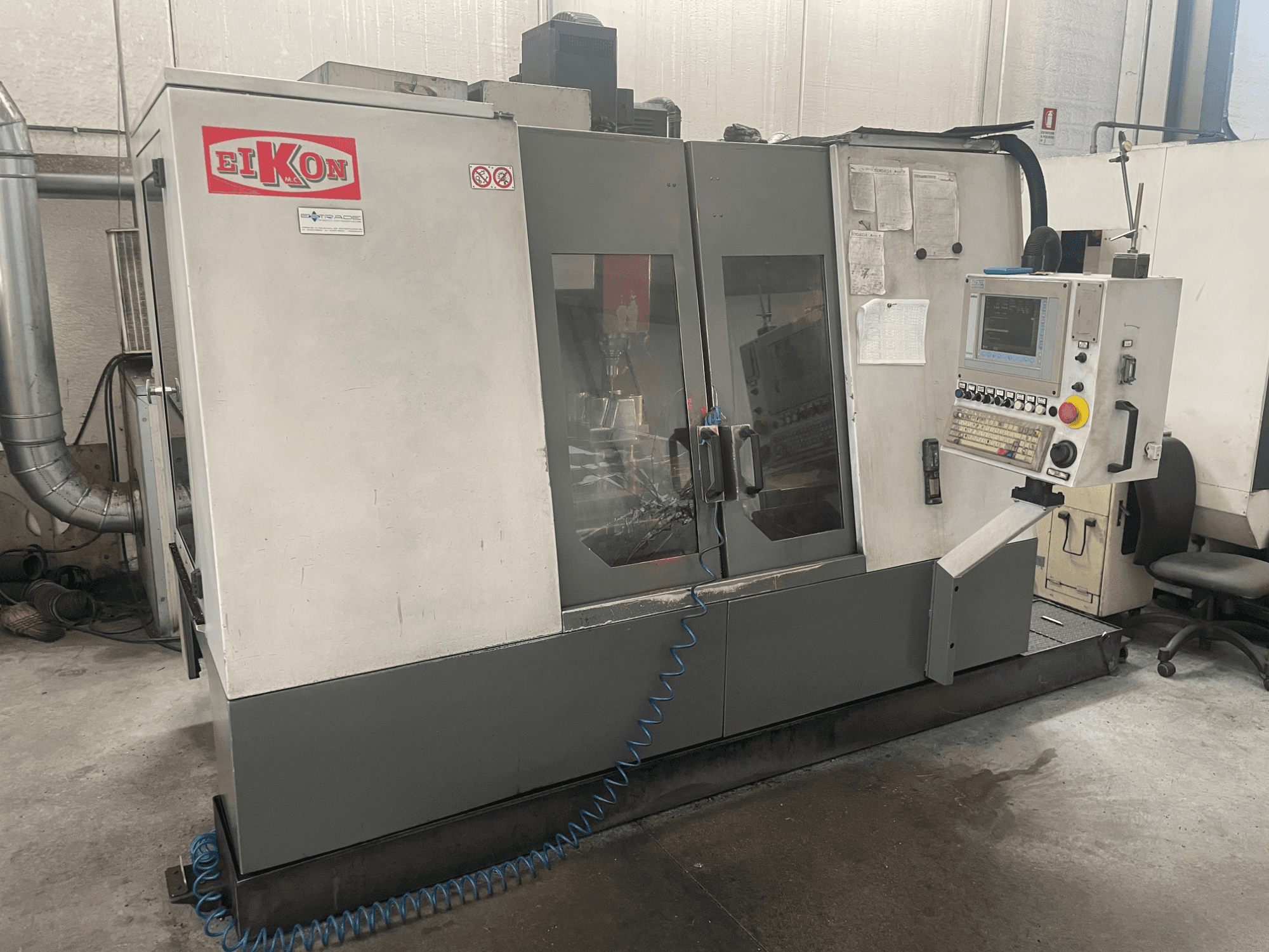 Máquina CNC EIKON vista de frente, con un panel de control, grandes puertas de cristal y una manguera de aire azul en el suelo.