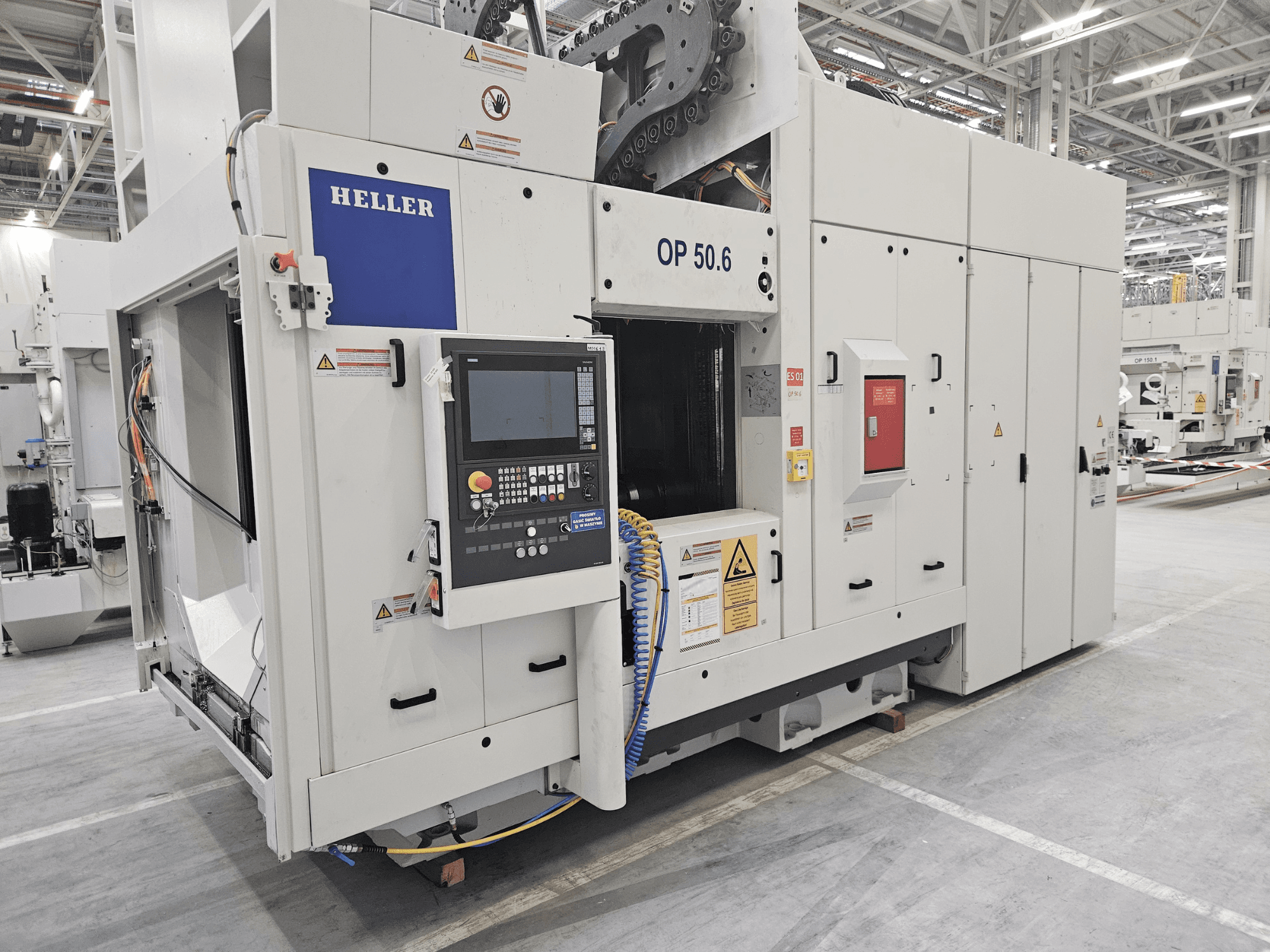 Centro de mecanizado Heller OP 50.6, vista frontal, con un panel de control, etiquetas de advertencia y mangueras visibles en un entorno de fábrica.
