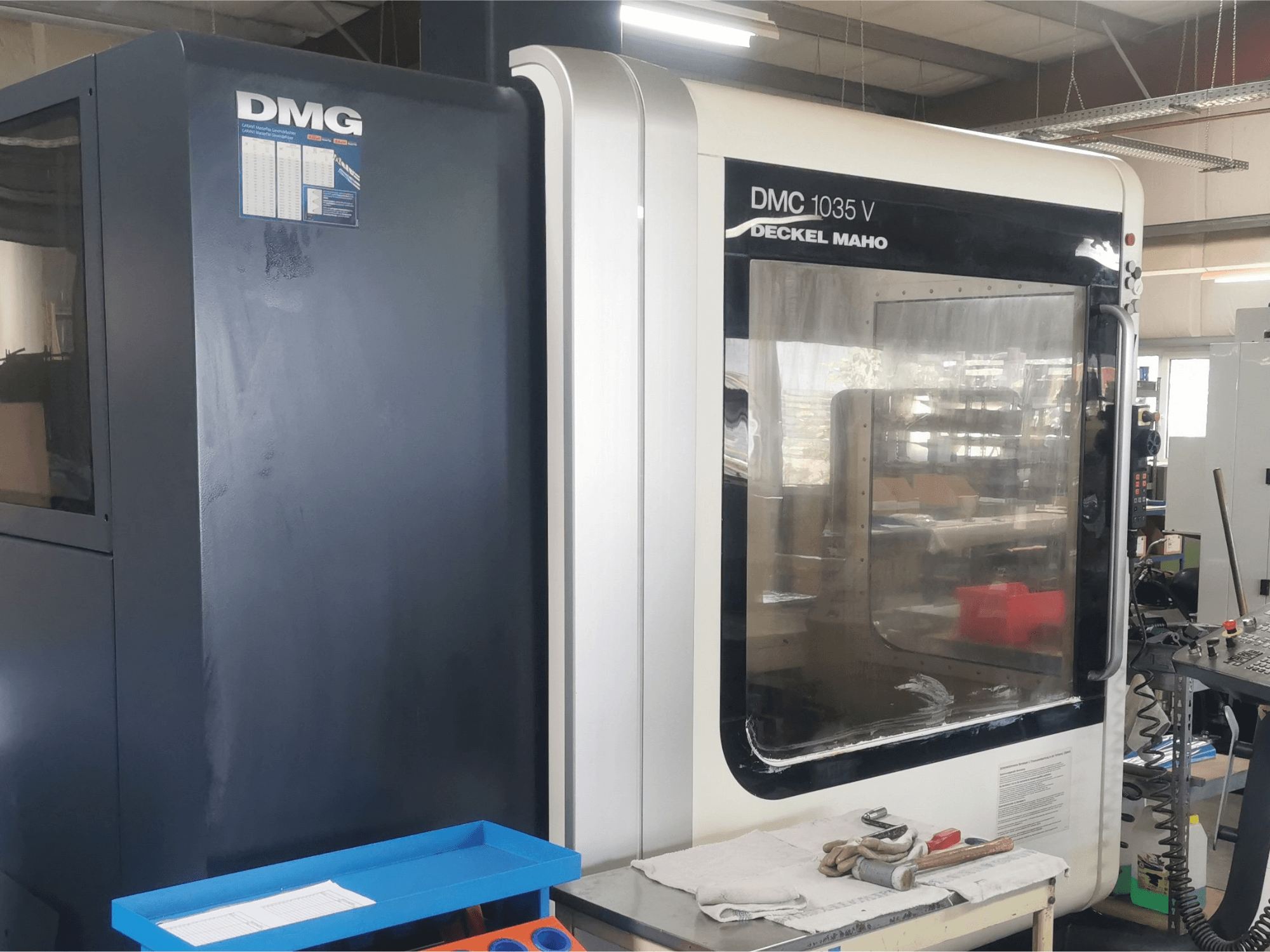 Fresadora DMG DMC 1035 V en un taller, vista frontal con bandeja de herramientas azul y guantes en la superficie.