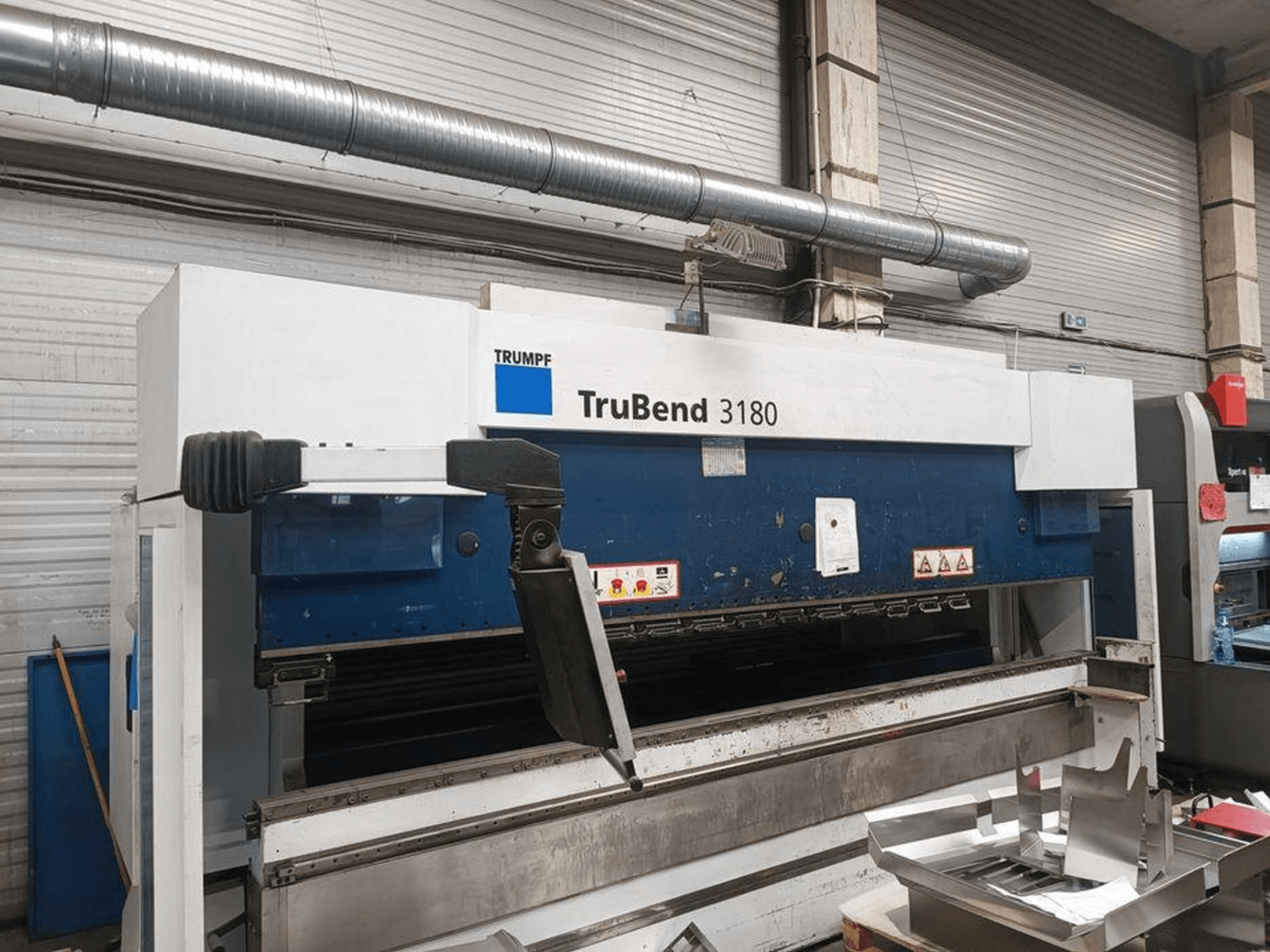 Plegadora Trumpf TruBend 3180 en un taller, vista lateral mostrando el panel de control y las piezas metálicas sobre la mesa.
