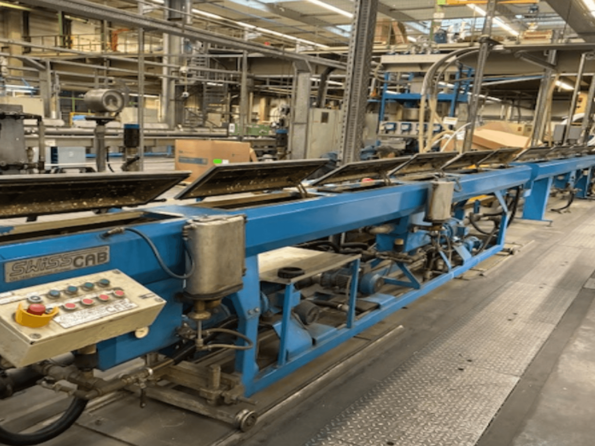 Máquina industrial SwissCab azul en un entorno de fábrica, con botones de funcionamiento y un sistema de transporte, vista lateral.