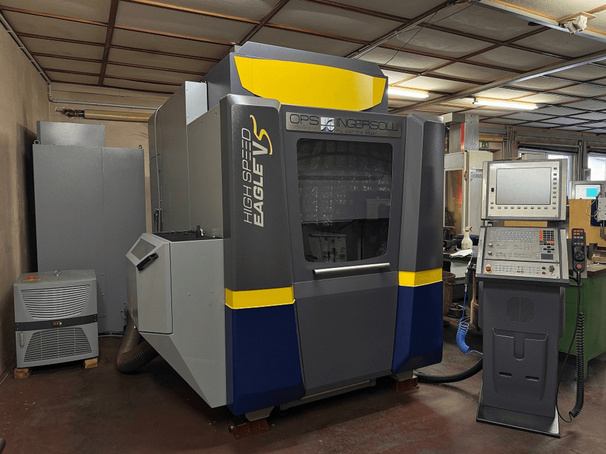 Máquina Eagle V de alta velocidad de OPS Ingersoll, vista lateral, con panel de control y equipos adyacentes en un entorno industrial.