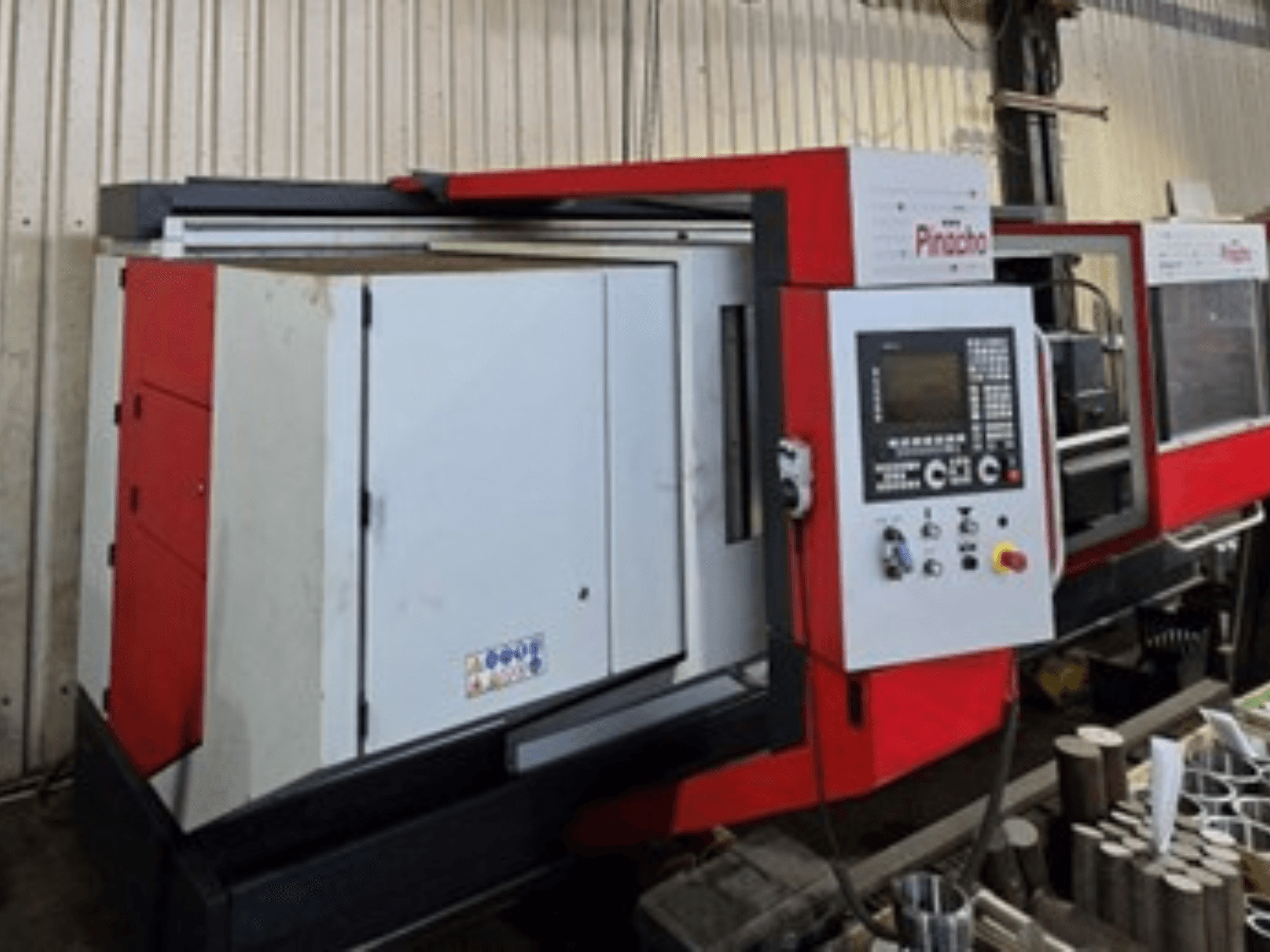 Máquina CNC Pinacho en rojo y blanco, vista lateral, con panel de control y materiales en el suelo, mostrando la configuración operativa.