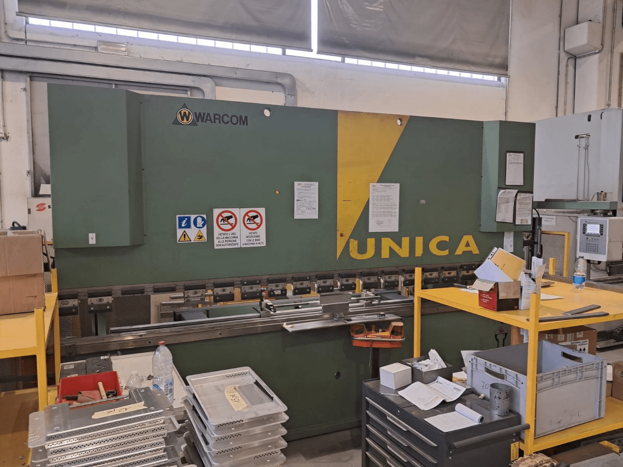Prensa hidráulica Warcom UNICA 100-40, vista frontal, cuerpo verde con detalles en amarillo, señales de seguridad visibles, herramientas y materiales sobre la mesa.